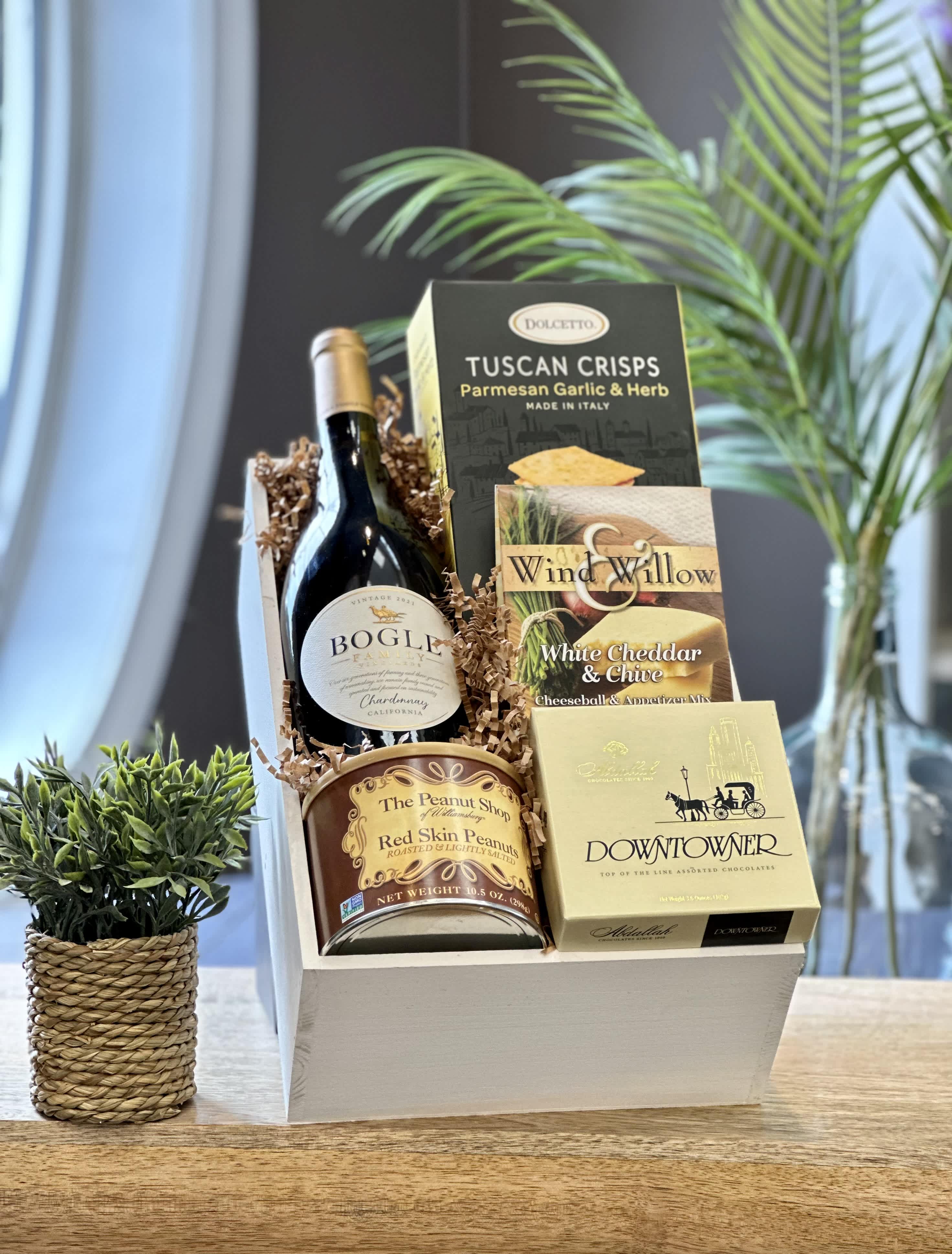 Gourmet gift crate with wine, crackers, peanuts, chocolates, and snack mixes on a wooden surface