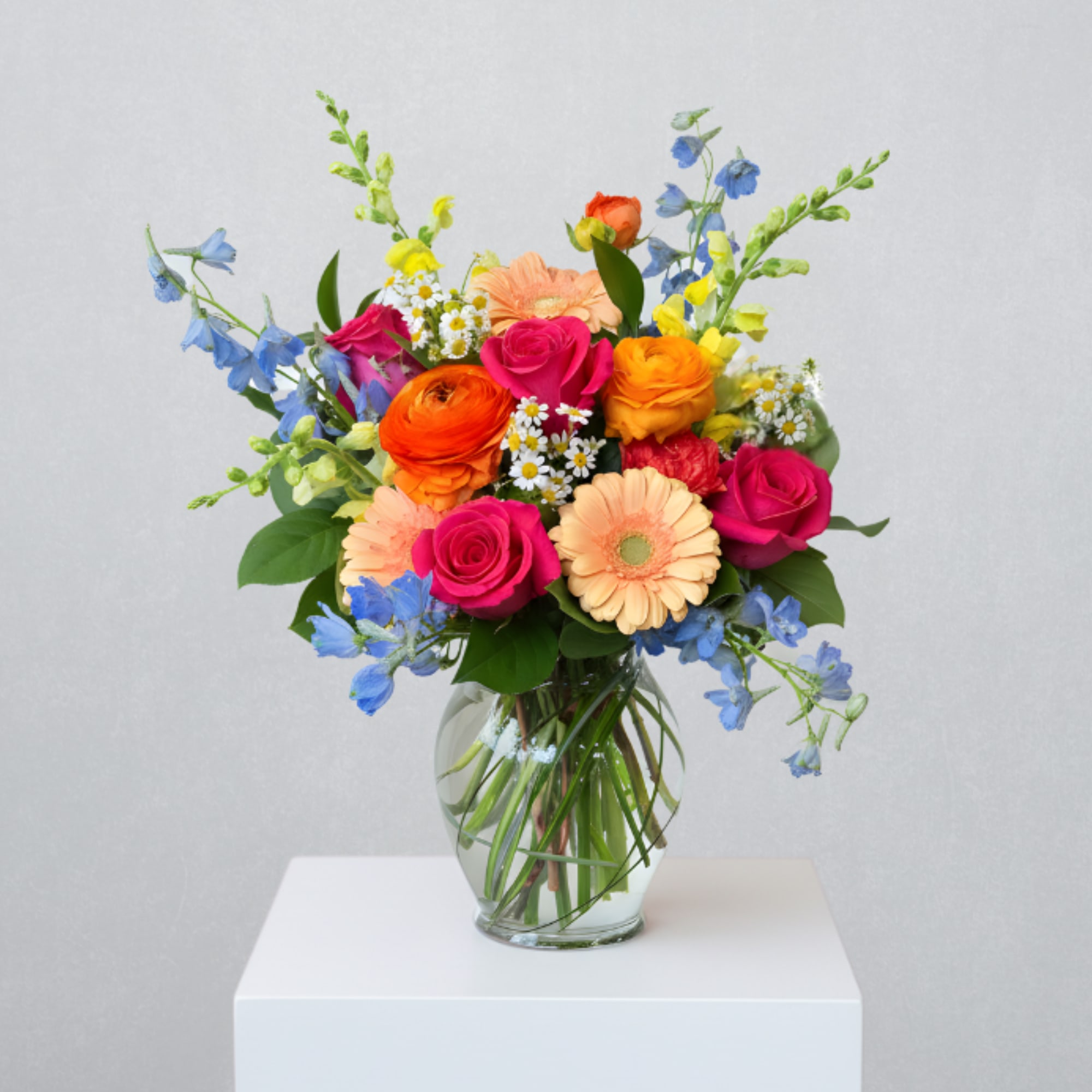 Bright mixed bouquet of pink roses, orange blooms and blue flowers in a clear glass vase.