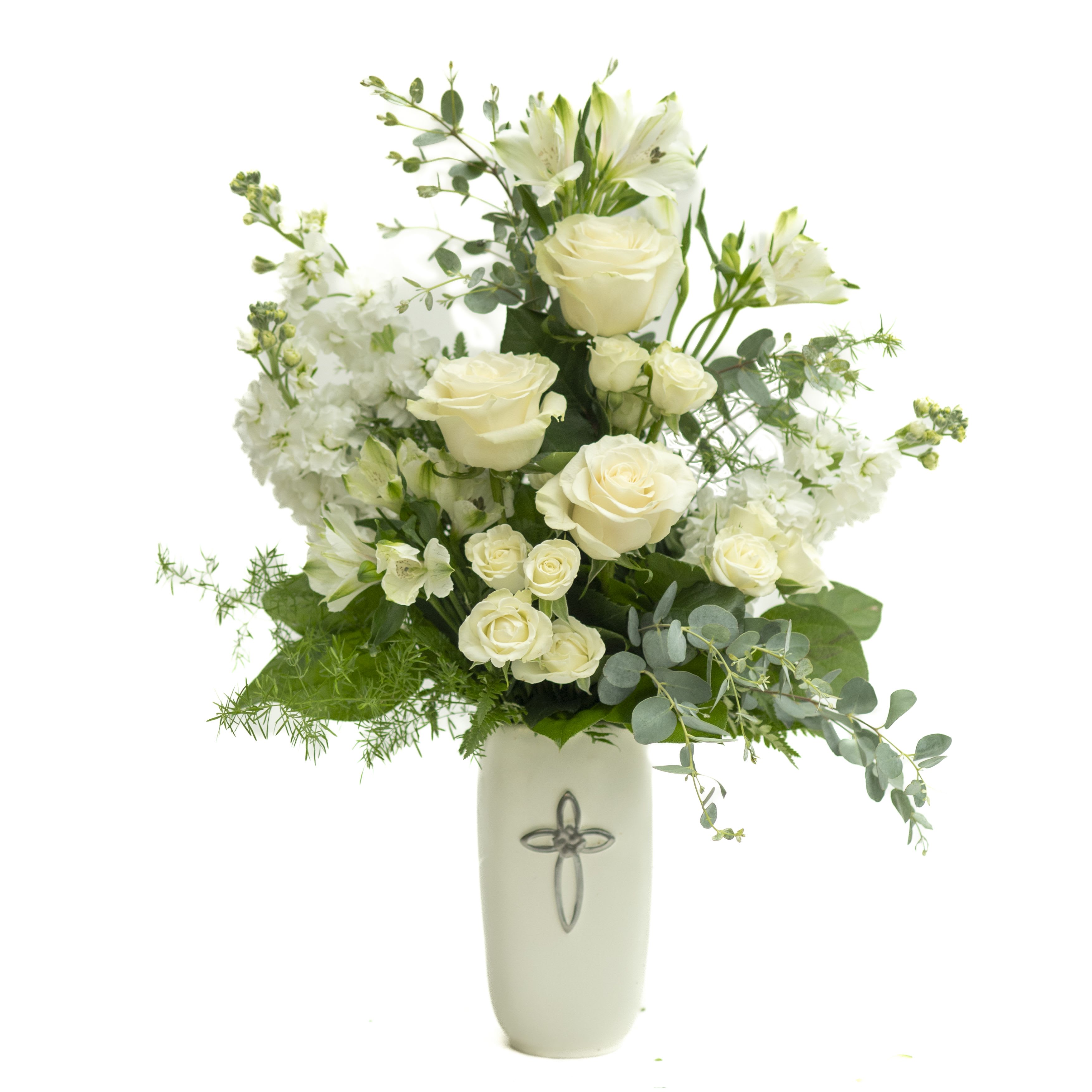 Blessed - Pure white roses and lilies rise from a tall cream ceramic vase embossed with a subtle cross, creating a serene sympathy arrangement that gently says "thinking of you." The upright, airy silhouette and soft white palette give this bouquet a formal, reverent presence perfect for funerals, memorial services, or sending comfort to someone grieving; it also works as a quiet, respectful gift for a church or spiritual occasion.  Clustered spray roses, alstroemeria, stock and delicate filler blooms are layered with varied green foliage and eucalyptus-like accents for texture and movement. The ceramic vase adds weight and permanence, so the arrangement sits tall and composed on a mantel, altar, or bedside table while the mixed greenery softens the overall look.  Designed to offer calm and consolation, this arrangement helps the recipient feel seen and supported during a difficult time or when you simply want to let them know you care. Delivered ready to display in its ceramic vessel; to keep blooms fresh, place in a cool spot away from direct sun and follow any care card included with the arrangement.