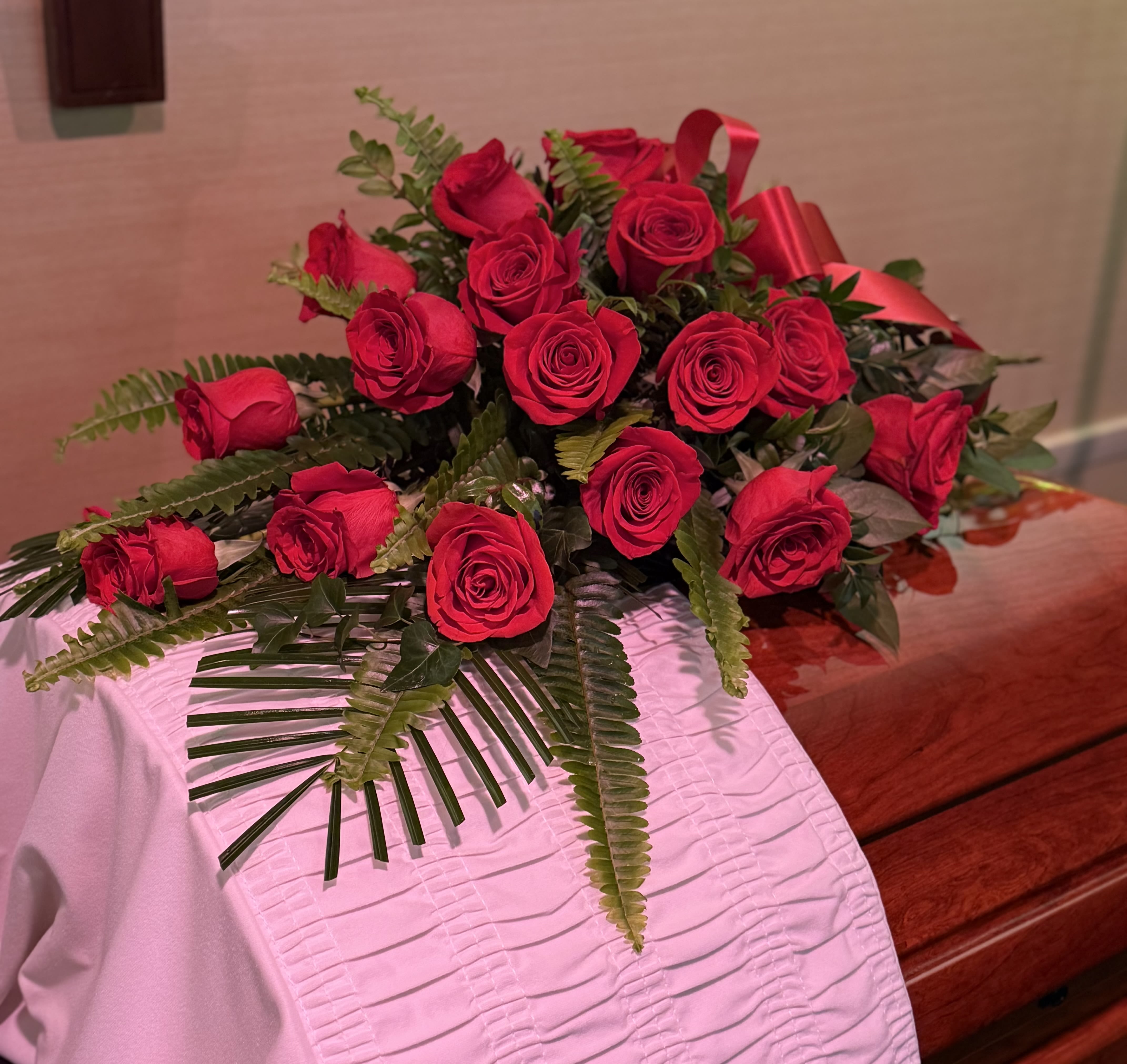 Red roses arranged with fern greenery and a red ribbon on a white cloth-covered casket