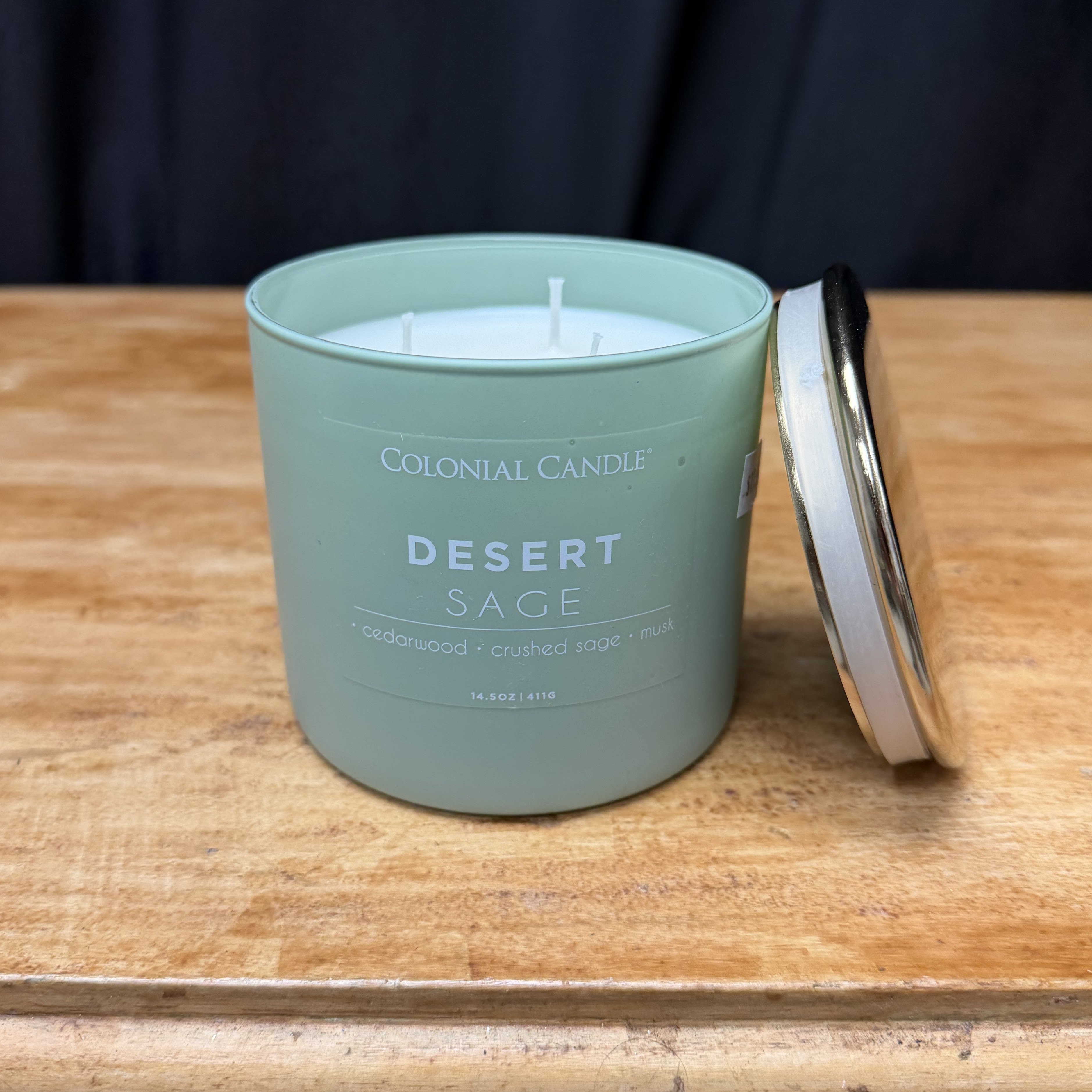A sage green candle in a metal tin with its lid beside it
