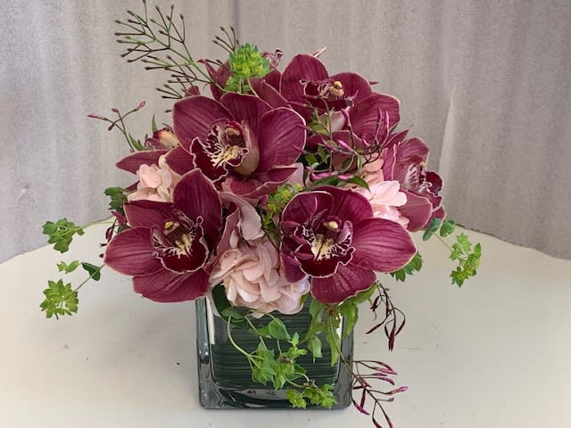 Deep purple orchids arranged in a square glass vase with pale pink blooms.