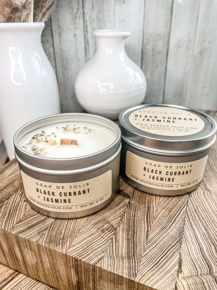 Two lidded tin candles labeled Black Currant + Jasmine on a wood block with white vases behind