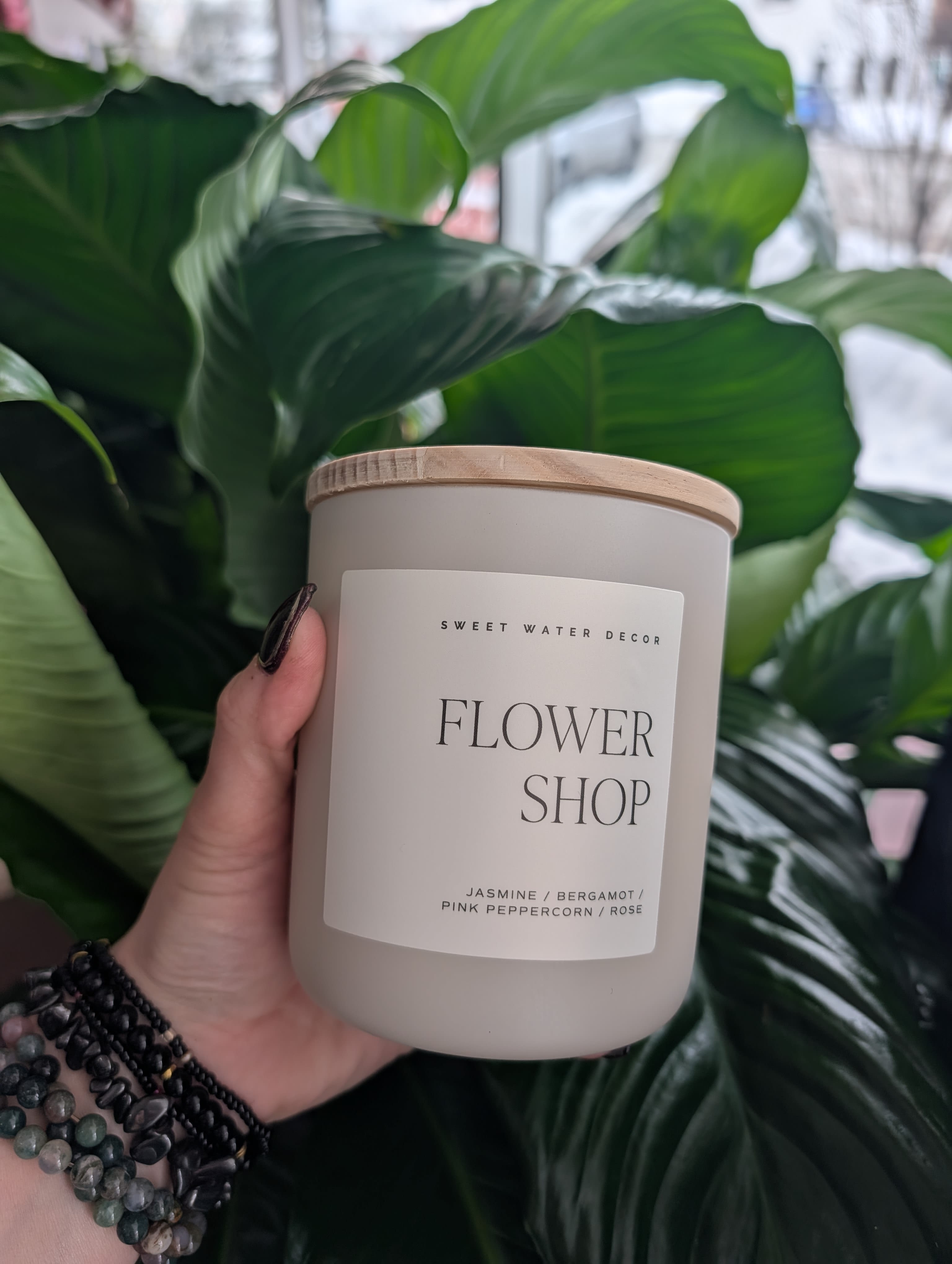 Hand holding a flower shop candle jar in front of large green leaves.