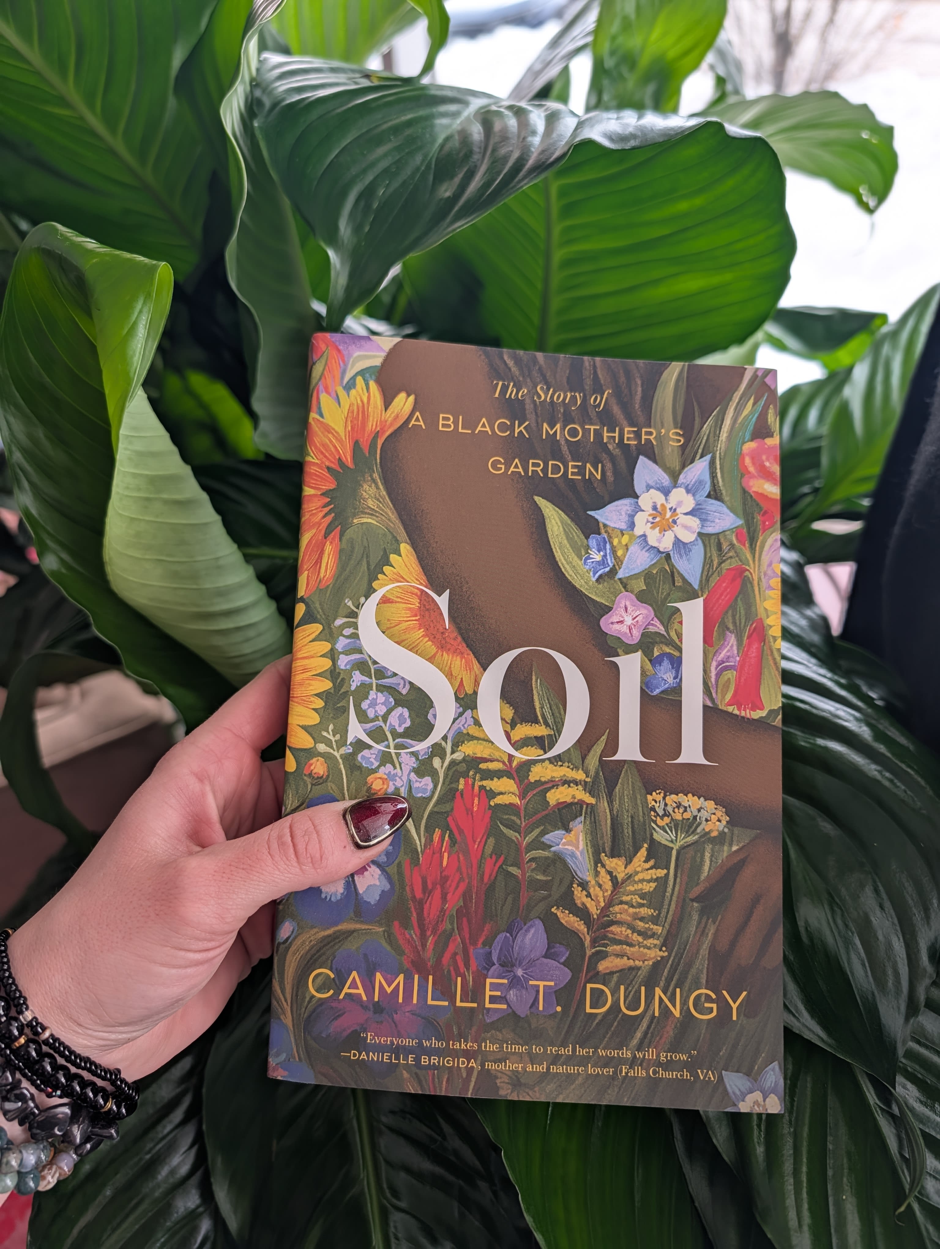Hand holding a floral-covered book in front of large green leaves.