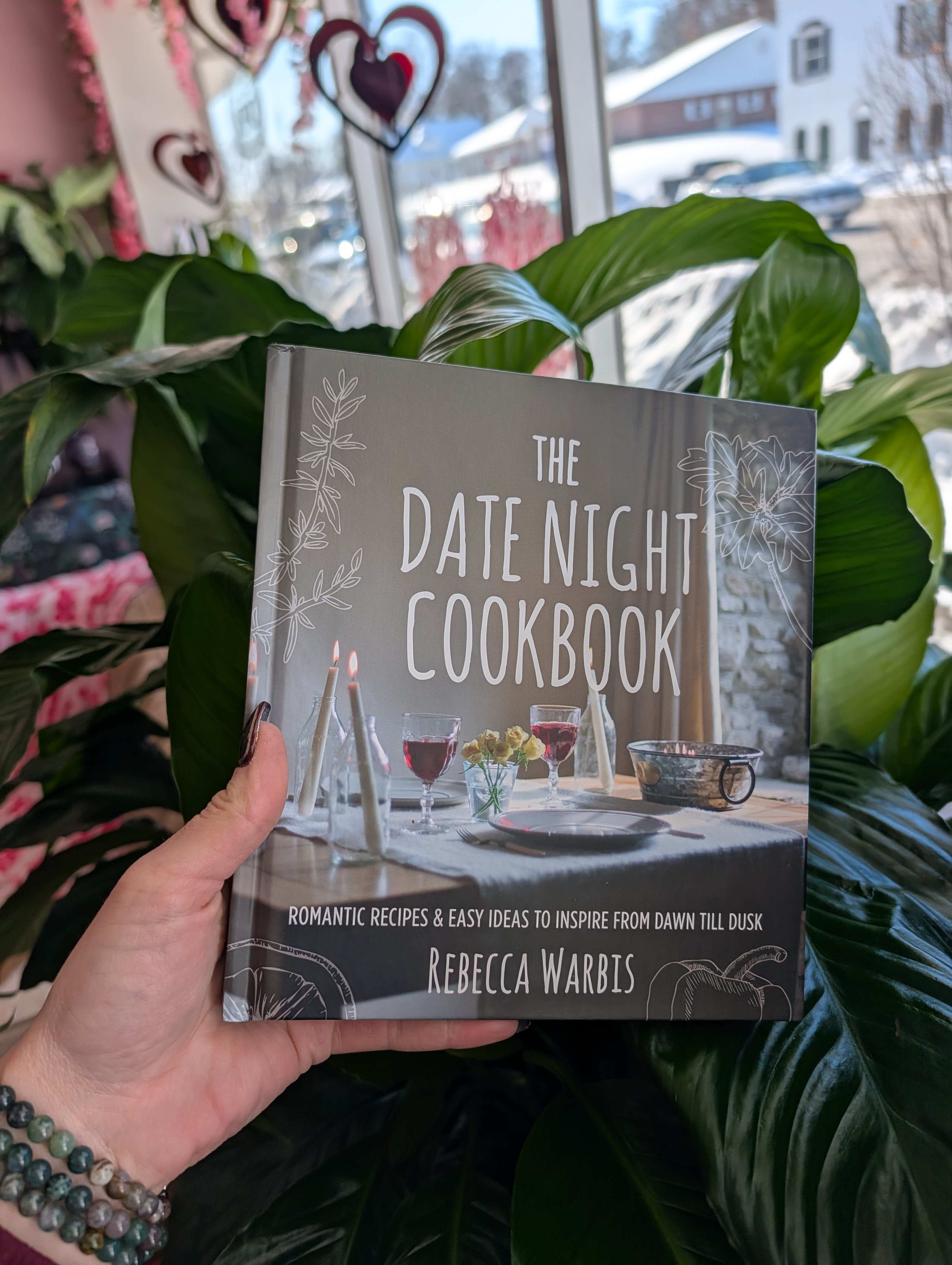 Hand holding a cookbook in front of large green houseplants
