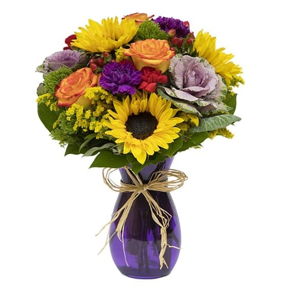 Sunflower Bliss - Somehow, almost magically, sunflowers can brighten a room...not to mention someone’s day! Sunflower beauty is front and center in this blissful Fall celebration, surrounded by carnations, green trick, solidago, hypericum, roses and kale in a purple jordan vase with raffia tie. Arrangement Height 17", Length 13"