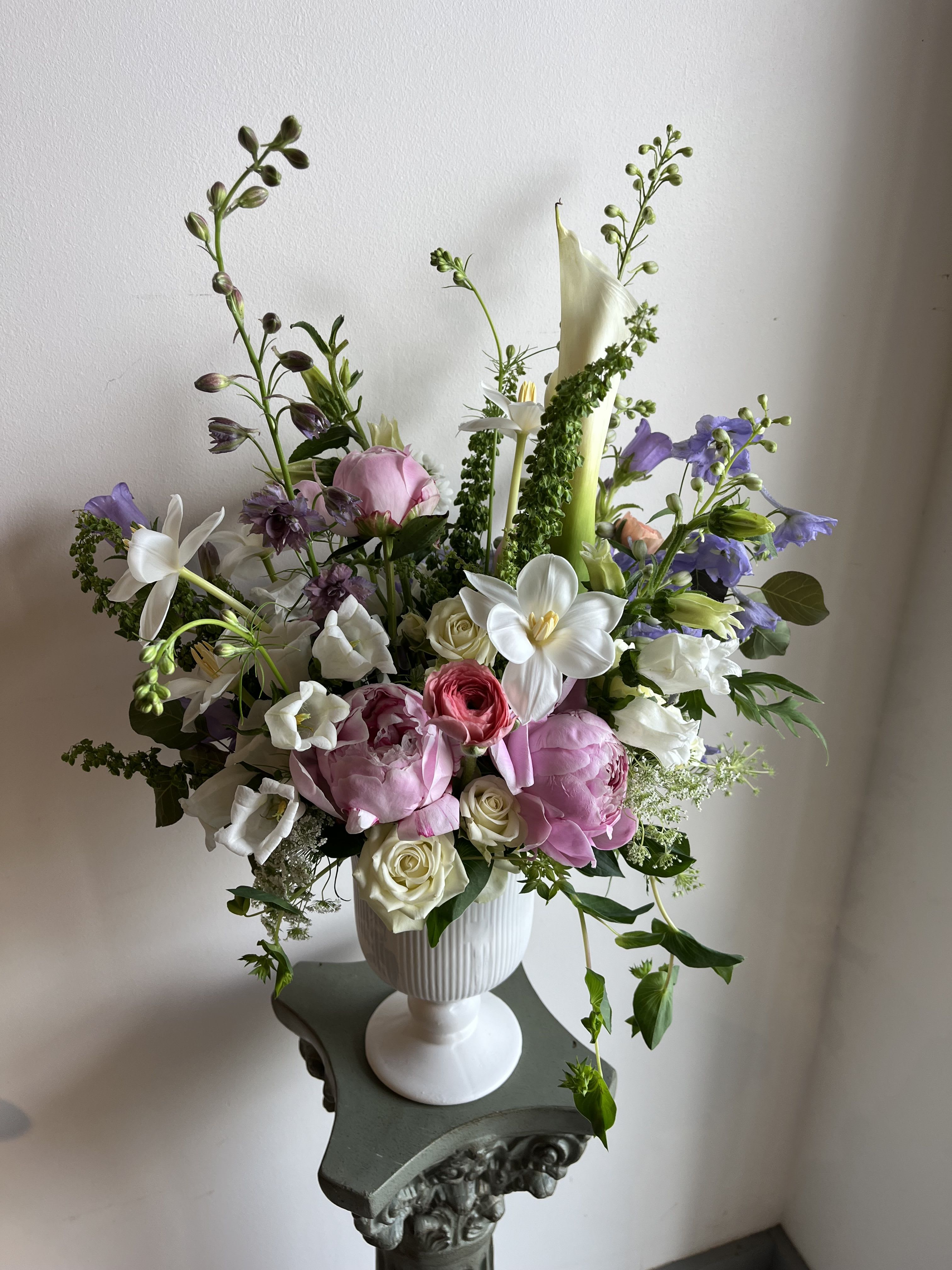 The Madeline - The Madeline is a soft and romantic garden-style floral arrangement designed to evoke timeless elegance and graceful beauty. Overflowing with lush blush pink peonies, creamy white roses, airy lavender delphinium, sculptural calla lilies, and whimsical seasonal blooms, this arrangement captures the feeling of a freshly gathered luxury garden bouquet. Designed in a classic white pedestal vase, The Madeline features an organic, asymmetrical silhouette with gentle movement and layered textures that feel both refined and effortlessly natural.  Its soft pastel palette and airy composition create a sense of warmth, femininity, and quiet luxury, making it a beautiful choice for weddings, anniversaries, sympathy gestures, baby showers, elegant events, or elevated everyday décor. Every stem is thoughtfully placed to create softness, dimension, and flowing movement, allowing each bloom to shine while blending harmoniously together.  Part of our luxury floral collection, The Madeline is perfect for those searching for romantic flower arrangements, premium fresh flower delivery, luxury peony arrangements, elegant centerpiece designs, and sophisticated garden-inspired florals with a modern yet timeless aesthetic.