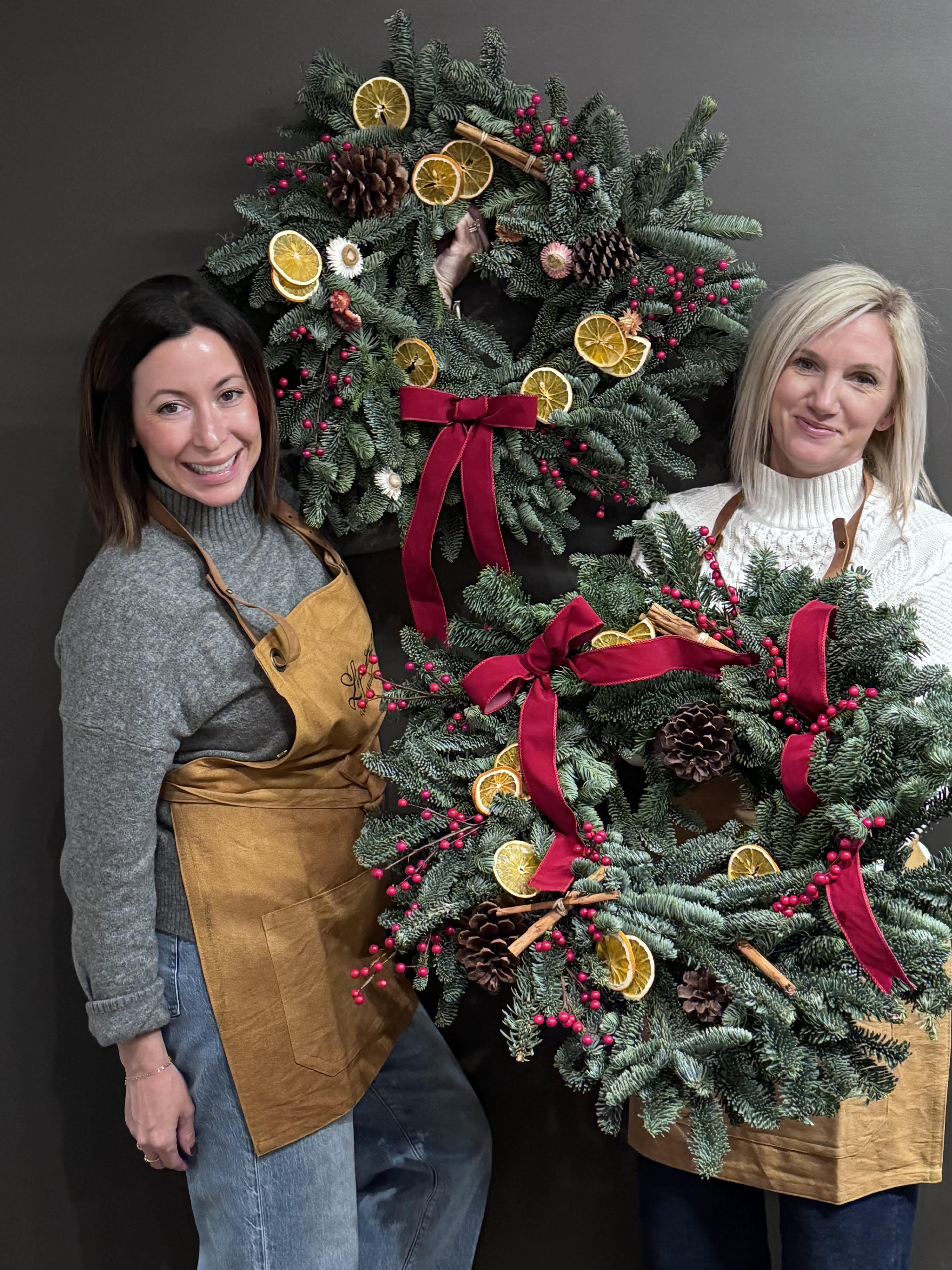 Two women hold decorated evergreen wreaths with red bows and dried citrus slices.