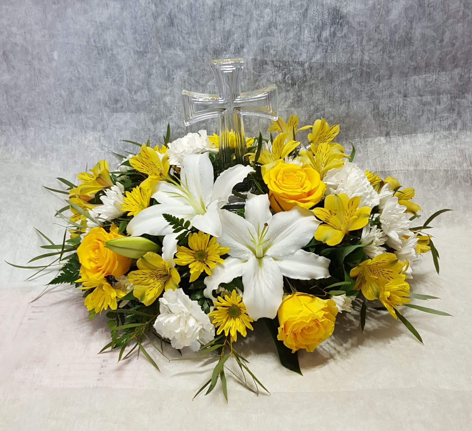 In My Savior's Arms - A fitting tribute to a life well lived, In My Savior's Arms reminds friends and family to rejoice in knowing their loved one is safe and truly at peace. This arrangement features a keepsake glass cross nestled among lilies, roses, daisies, cushion mums, and alstroemeria lily. COLORS AND FLOWERS WILL VARY WITH AVAILABILITY. Cross is 8.5" high.  Overall arrangement approximate size: 18"L x 12" H.  DELUXE version have more roses and lilies. This arrangement can be made in a variety of colors. PLEASE SPECIFY COLOR REQUESTS IN THE "Special Instructions" box.