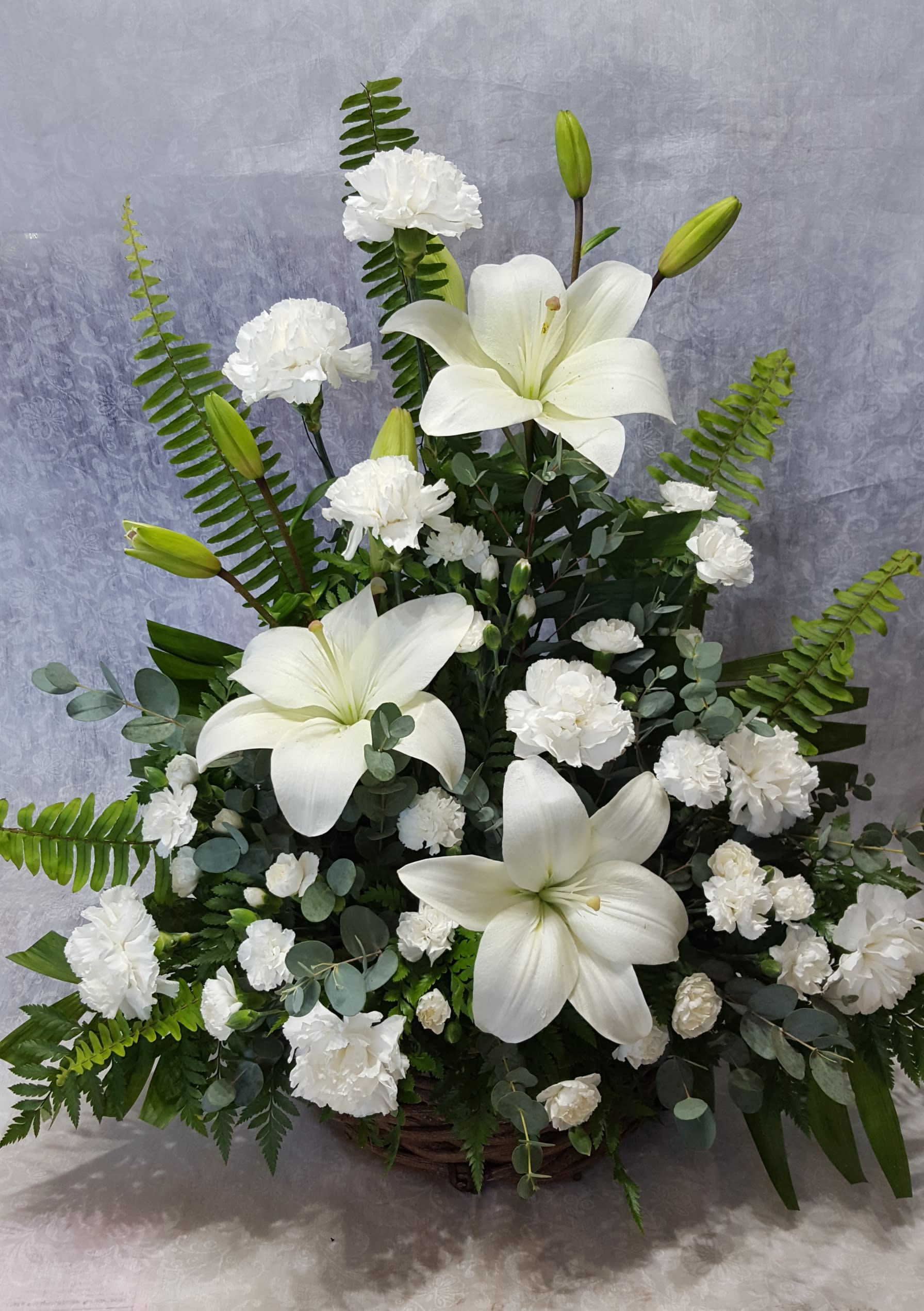 Lily Arrangement - Fresh white Asiatic lilies and carnations are mixed with rich greens such as salal and oregonia in a natural basket. A peaceful tribute for a beautiful life lived.  Approximate size: 24" H x 22"W
