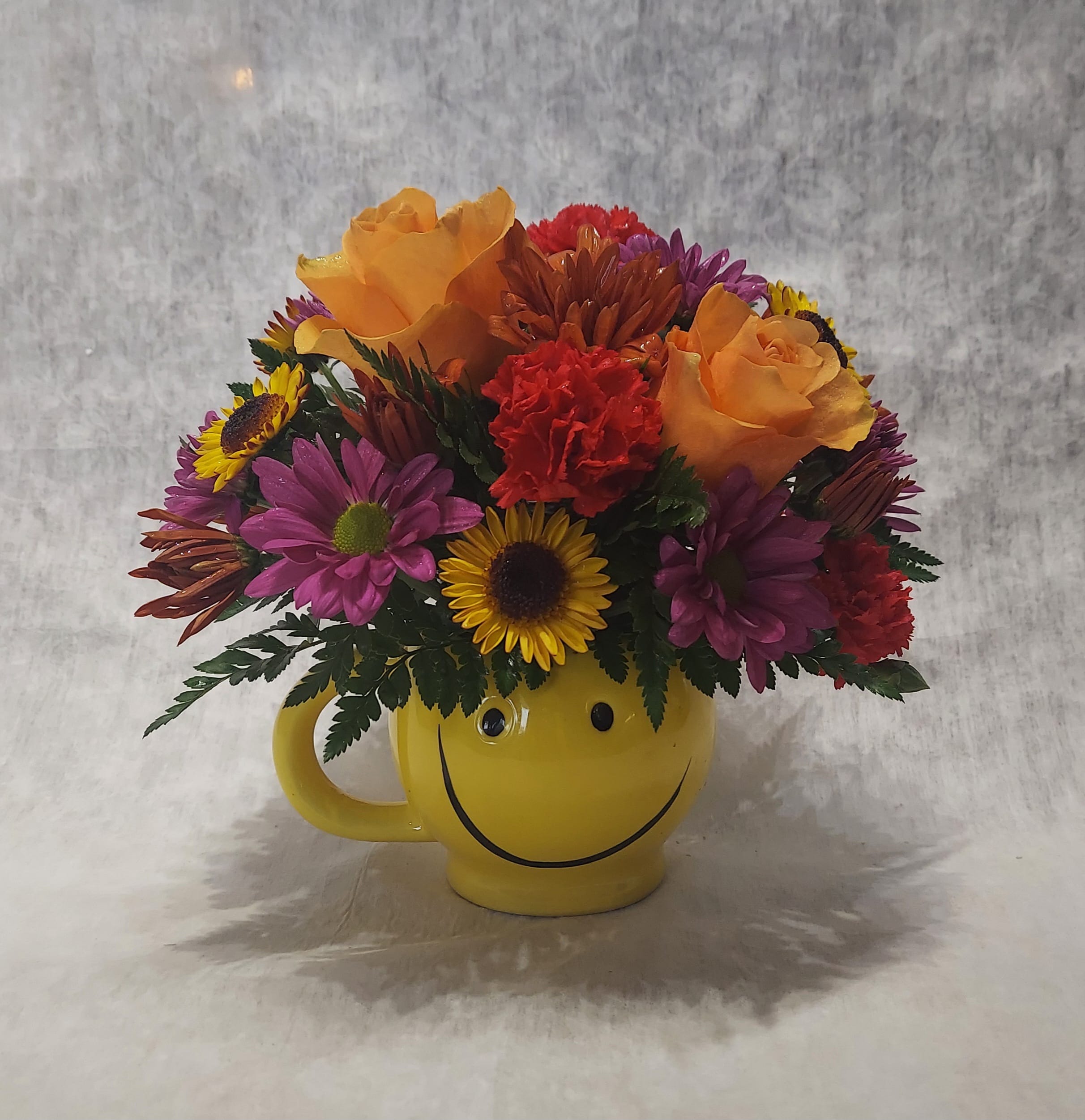 Autumn Smiles  - Brightly colored fall blooms make this smiley mug an Autumn favorite. Approximately 8.5" x 8.5"  COLORS OF FLOWERS WILL VARY WITH AVAILABILTY.