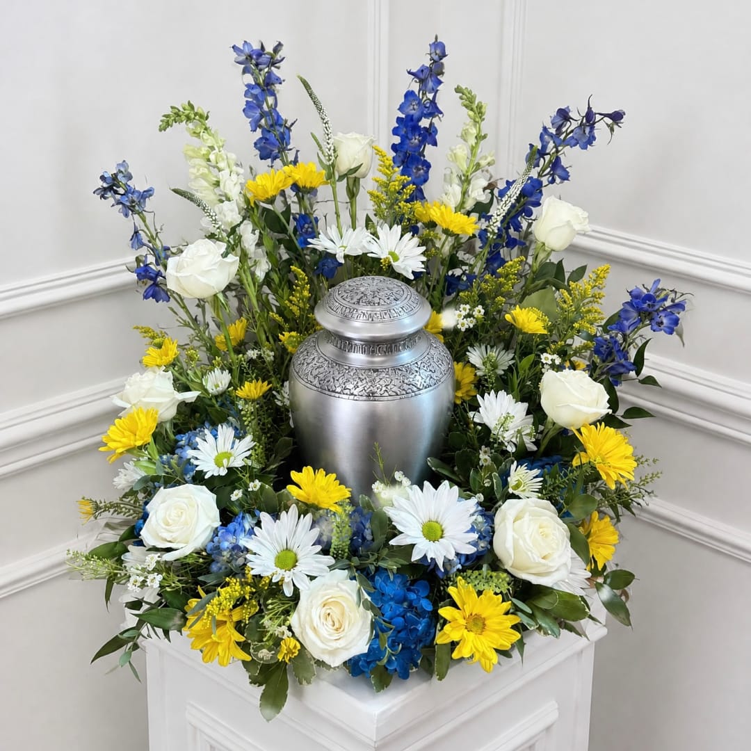 White roses, blue flowers, and yellow daisies arranged around a silver urn