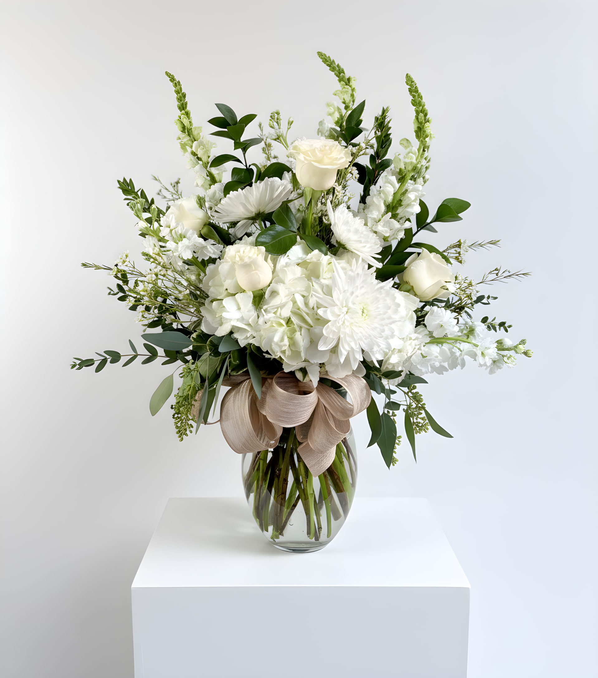 Tall white floral arrangement with roses and mums in a clear glass vase with a beige bow.