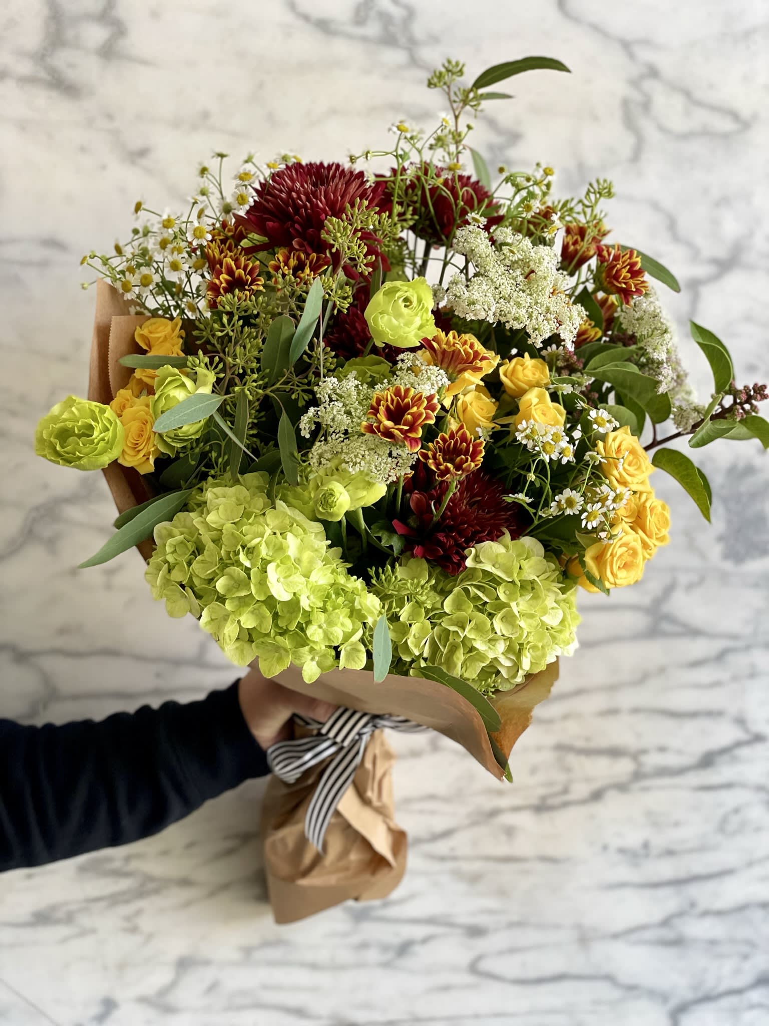 Mixed bouquet of yellow, lime green, and burgundy flowers wrapped in paper