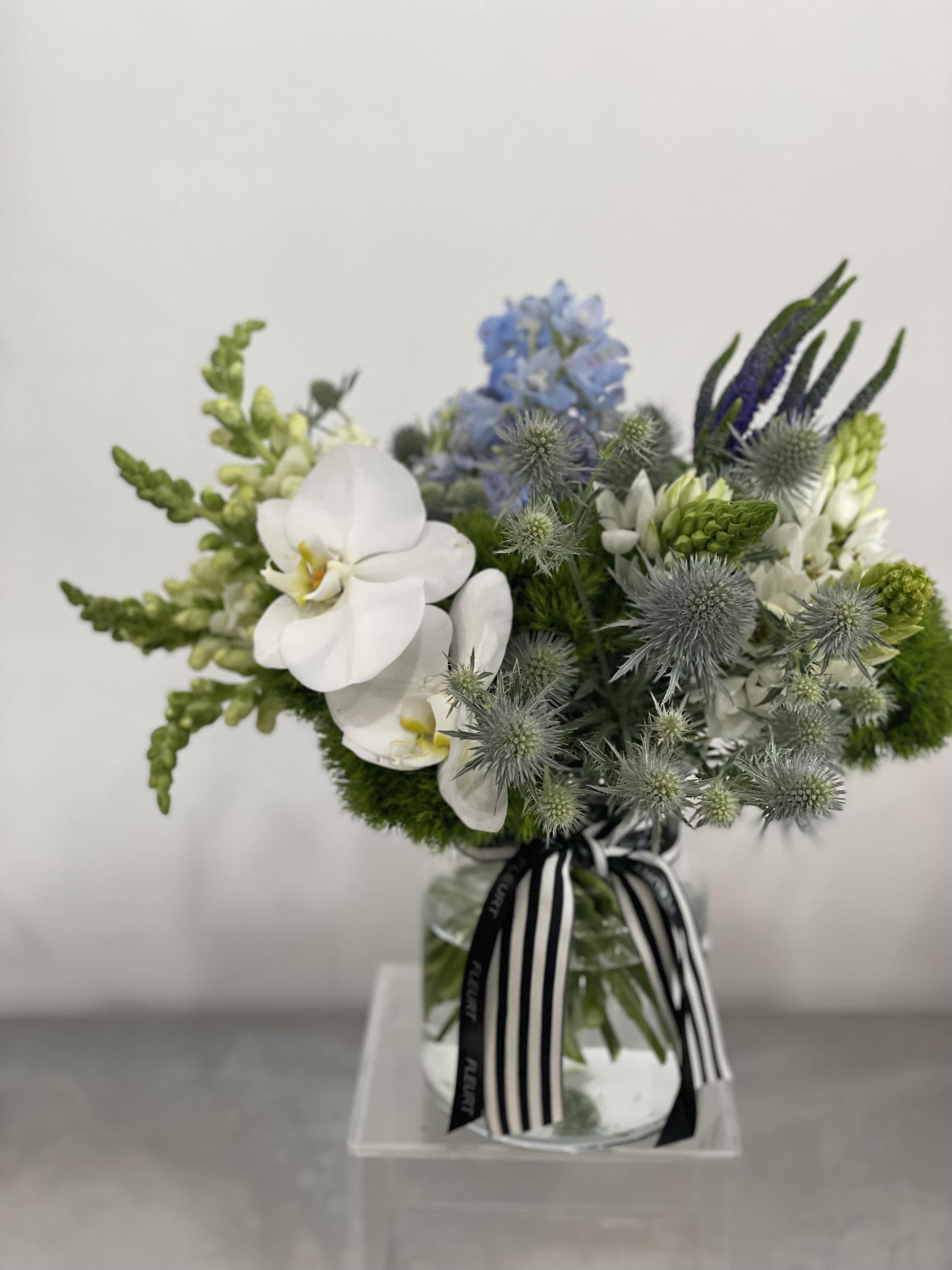 Low glass vase arrangement with white orchids, blue flowers, and green accents tied with a striped ribbon.