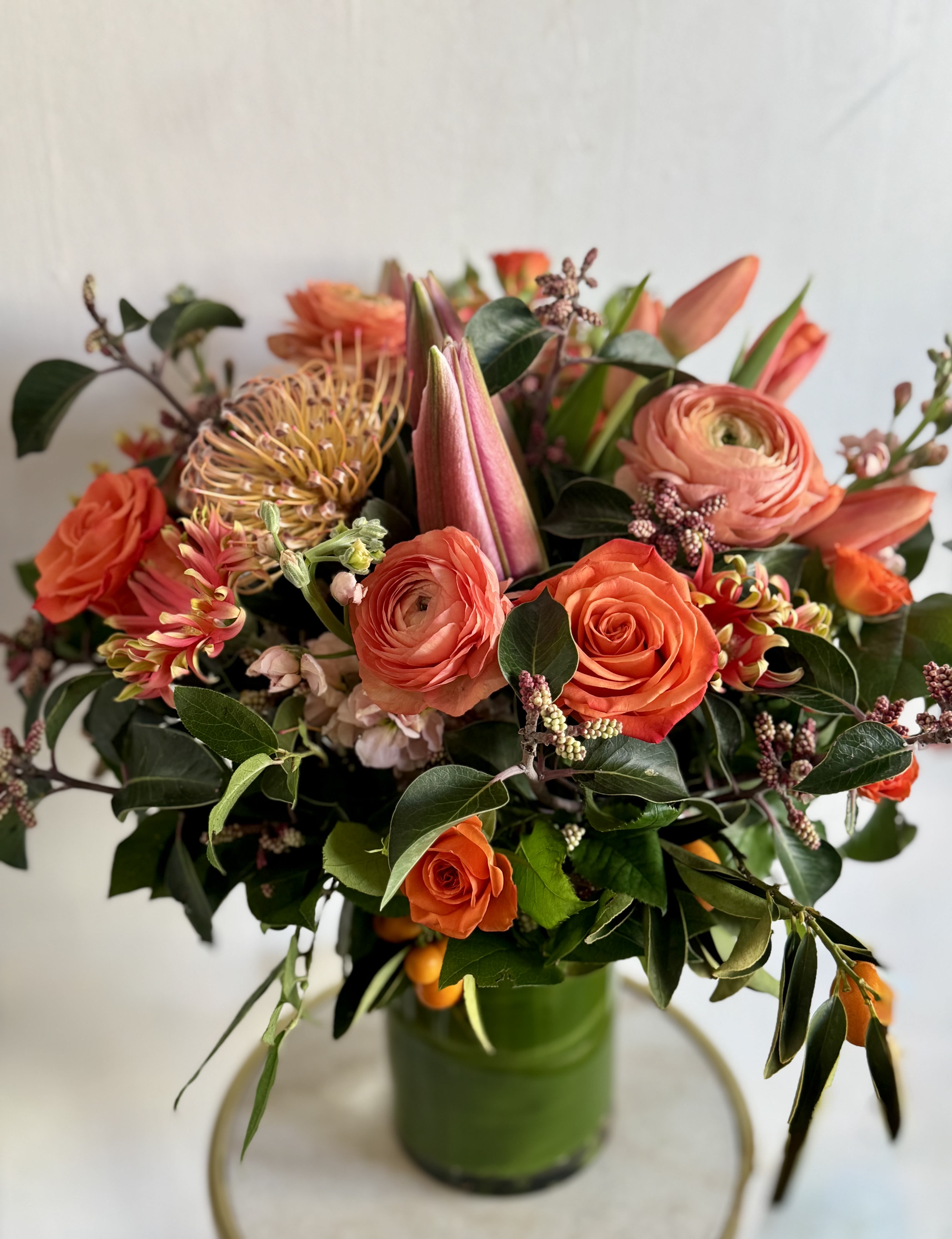 FANTA - Picture is to represent our work, may not look identical.  DESIGNERS CHOICE FLOWERS - ORANGE  + PEACH + YELLOW  are arranged densely in a clear glass vase, with dark glossy foliage accents and textured filler flowers lending a slightly wild, garden-style texture. The mix of soft peach and vibrant orange petals with touches of pink and green gives the design both warmth and fresh contrast; dimensions vary by size but the arrangement is intentionally full and rounded to feel generous from every angle. Vases may be substituted based on availability.  NOTES: We will choose the freshest and most interesting ingredients at the market to make this arrangement shine. Vessels may be substituted depending on availability.  If any design element such as color or flower/foliage are very important to your order please contact us to ensure availability first by calling the shop at 415 751 2747