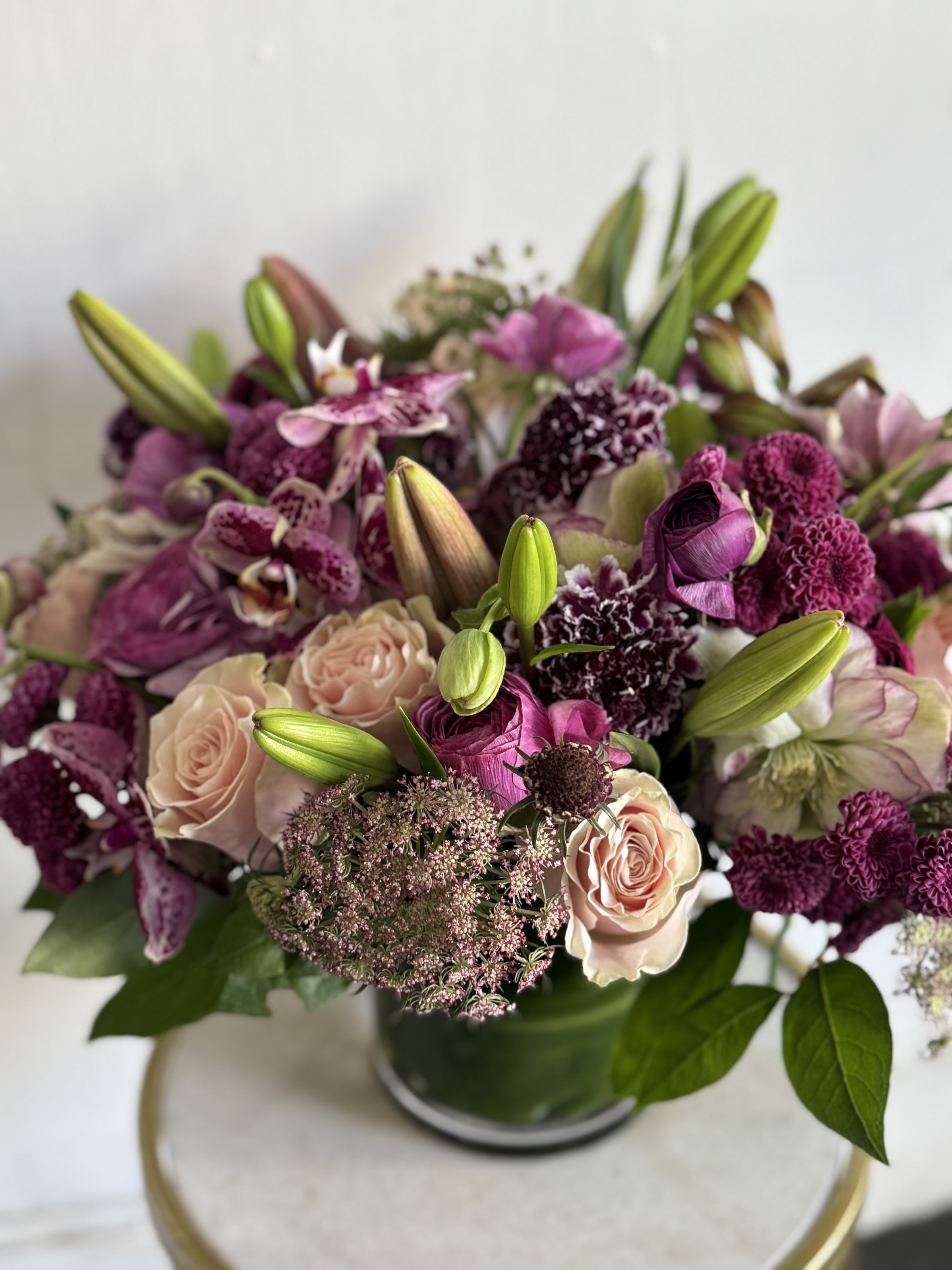 MOODY  - A lush, garden-style bouquet that reads romantic and refined, MOODY brings together blush roses, deep magenta ranunculus and plum-toned blooms with white and green accents. Perfect for Mother's Day or an anniversary when you want a softer, more intimate expression.  Built in a clear glass vase, this full arrangement layers textured petals, velvety roses, speckled orchid-like blooms, clustered dianthus and lush greenery-for a dense, tactile effect that reads both lush and composed. The palette of pink, purple, white and green creates striking contrast between soft peach-pink and rich berry and plum tones, with lily buds reaching above the main mass for elegance and scent. Vases may be substituted based on availability; we select the freshest, most interesting market ingredients to recreate this look for the recipient.  