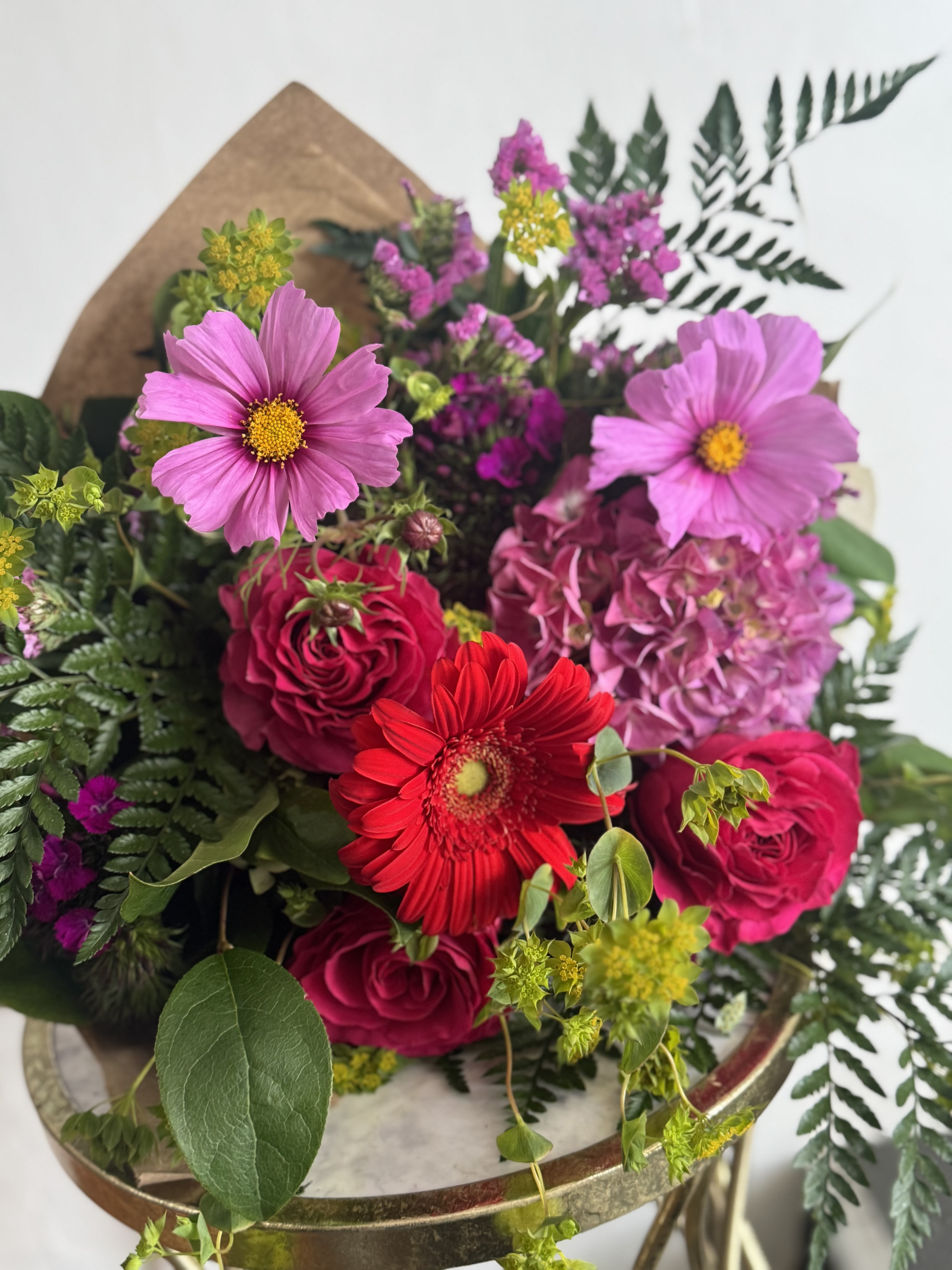 Berry Tones  - Designers Choice- Lovely bunch of berry toned flowers. Designers Choice may include all or most of the items pictured, but will feature the same general color and composition.   This mixed bouquet layers airy, cosmos-like blooms, classic roses, hydrangeas, and daisies with pops of small yellow-green accents and varied foliage for texture. Hand-tied in brown kraft paper for a natural, gift-ready presentation, the arrangement feels full and loose rather than tightly formal, giving it a fresh-from-the-garden movement and scent.  Sizes are available in a hand-tied bouquet wrap; care is simple-trim stems, refresh water, and keep away from direct heat to prolong the blooms.