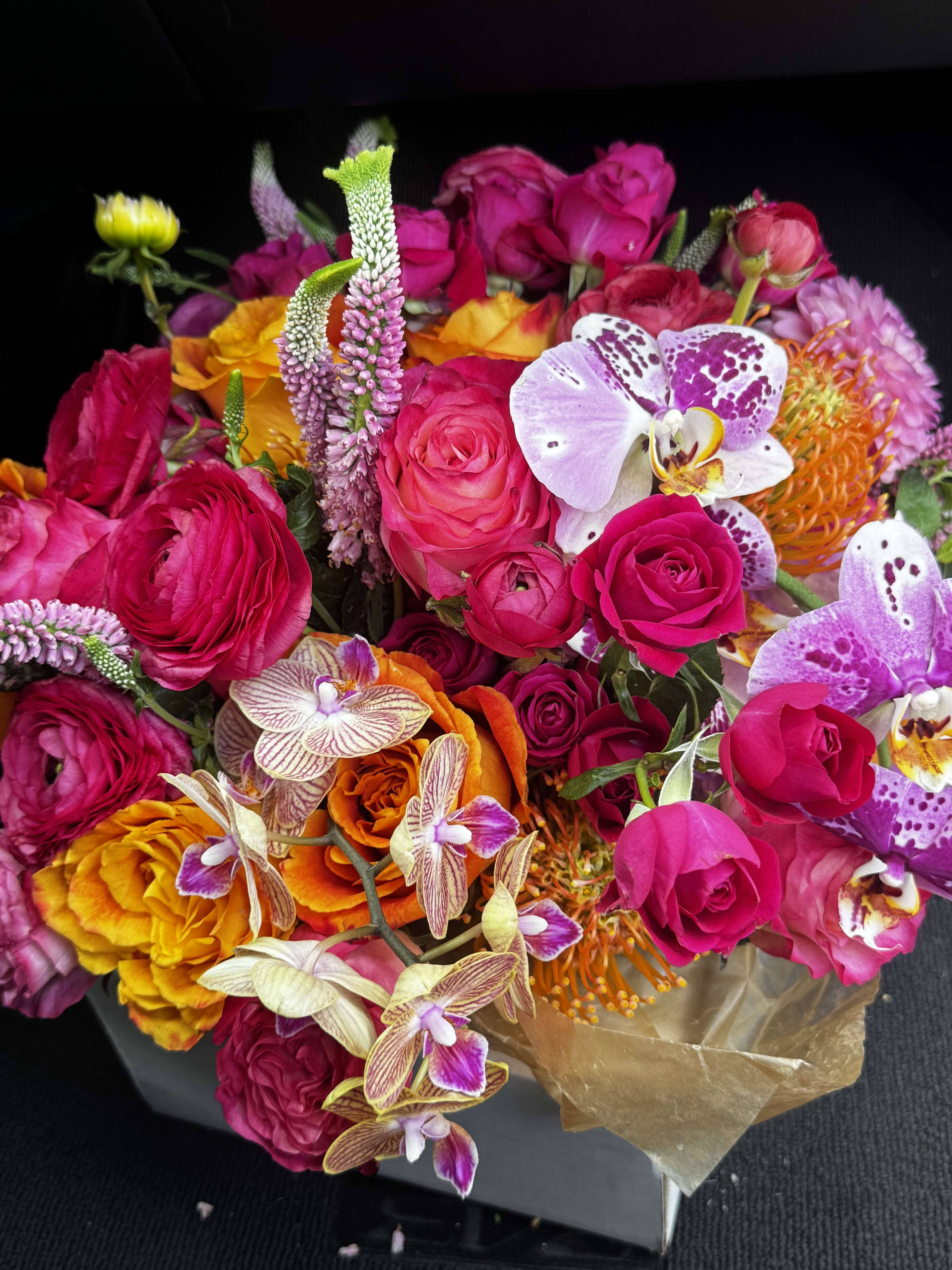 Bright mixed bouquet of pink roses, orchids, and orange blooms in brown wrap