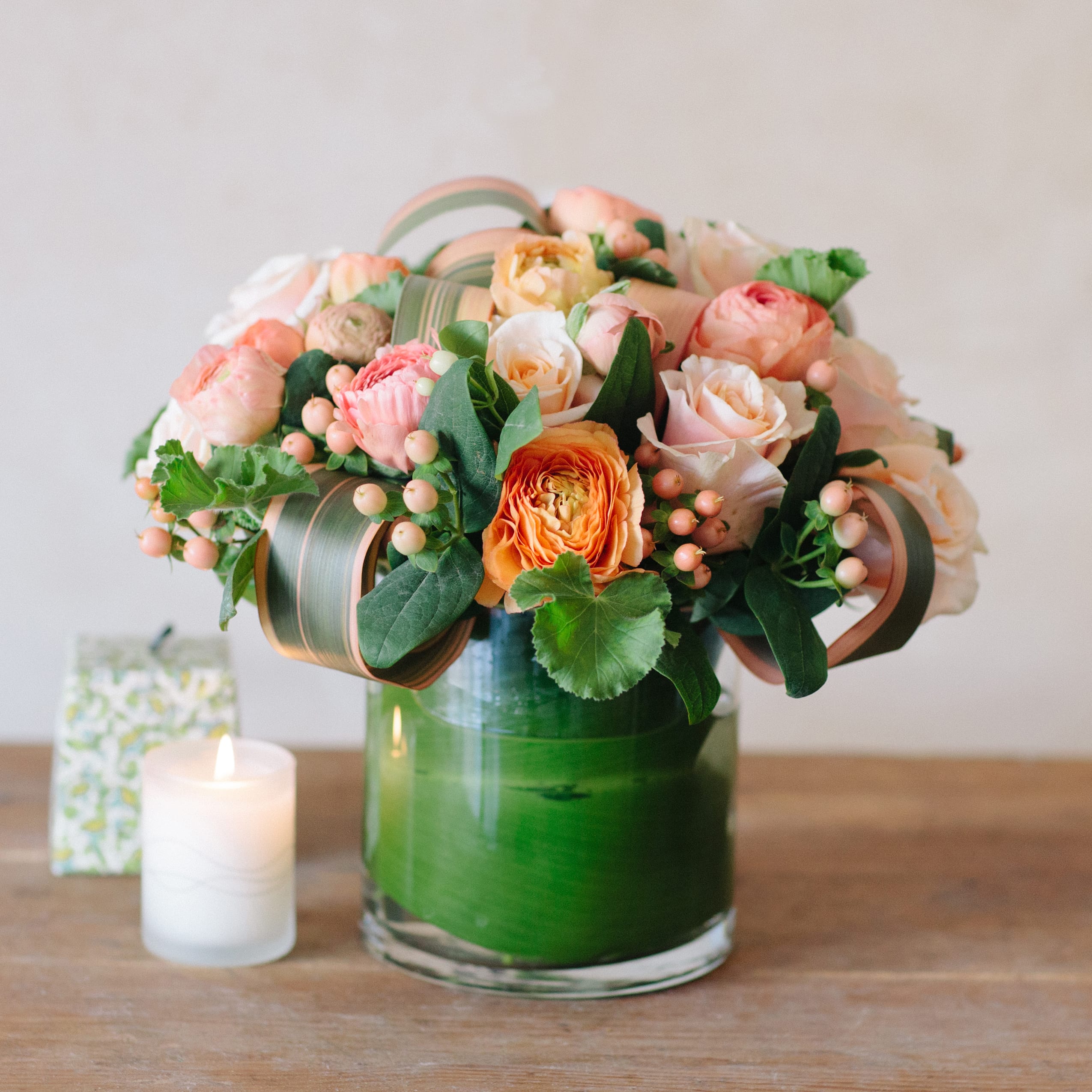 Short round arrangement of peach and cream roses in a clear glass vase with green accents