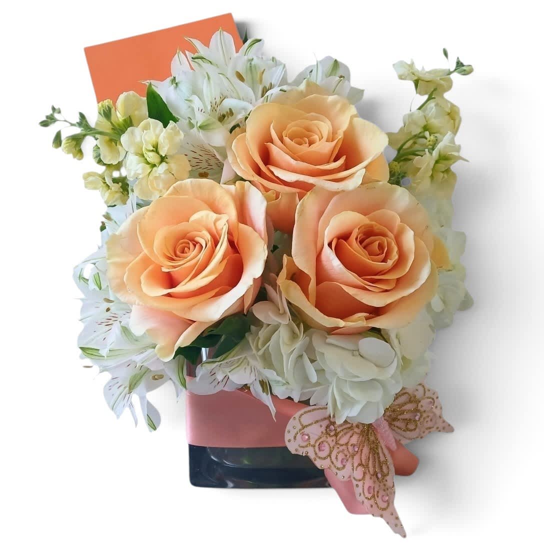 Peach roses and white flowers arranged in a square vase with a pink ribbon