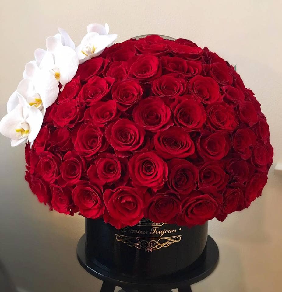 Round hatbox of densely packed red roses with a side accent of white orchids in a black container.