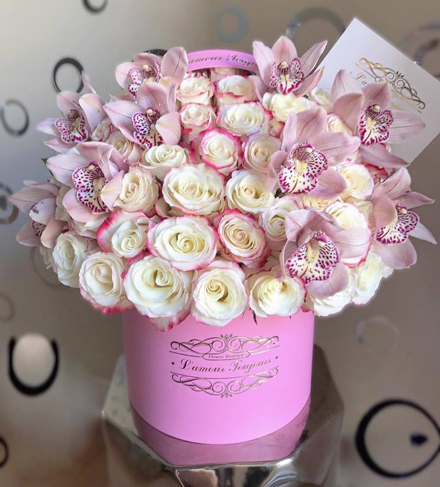 Round pink hat box filled with cream roses edged in pink and pale pink orchids, with a small card tucked in.