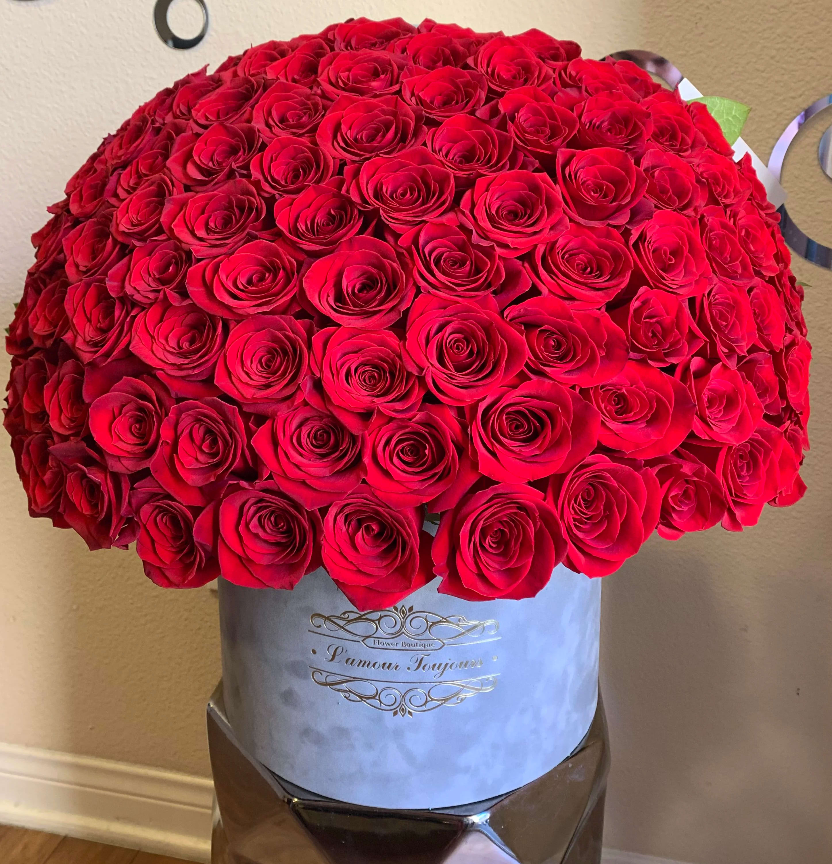 100 Red Roses Signature Box - A dramatic dome of 100 red roses forms a flawless canopy of blooms - the kind of signature bouquet that makes someone feel truly adored. Perfect as an anniversary or Valentine's Day statement, this luxury rose arrangement also reads as an unforgettable birthday, Mother's Day, or congratulations gift: the intense crimson and seamless round shape create a powerful impression of love, celebration, and admiration.  Each long-stem rose is tightly clustered into a velvet-like dome and presented in a pale gray hatbox-style container with a gold printed label, an elevated, gift-ready display. lt turns the piece into a striking centerpiece for weddings, graduations, corporate gifts, or sympathy and funeral settings when solace and beauty are needed.  This is the kind of arrangement that makes the recipient feel seen - whether you're sending a "just because" surprise, saying "I'm sorry," celebrating a new baby or prom, or honoring a milestone for Dad on Father's Day. Sized to impress and easy to place on a table or mantle, the bouquet arrives ready to display; for best longevity, keep in a cool spot and add fresh water as recommended with boxed arrangements.