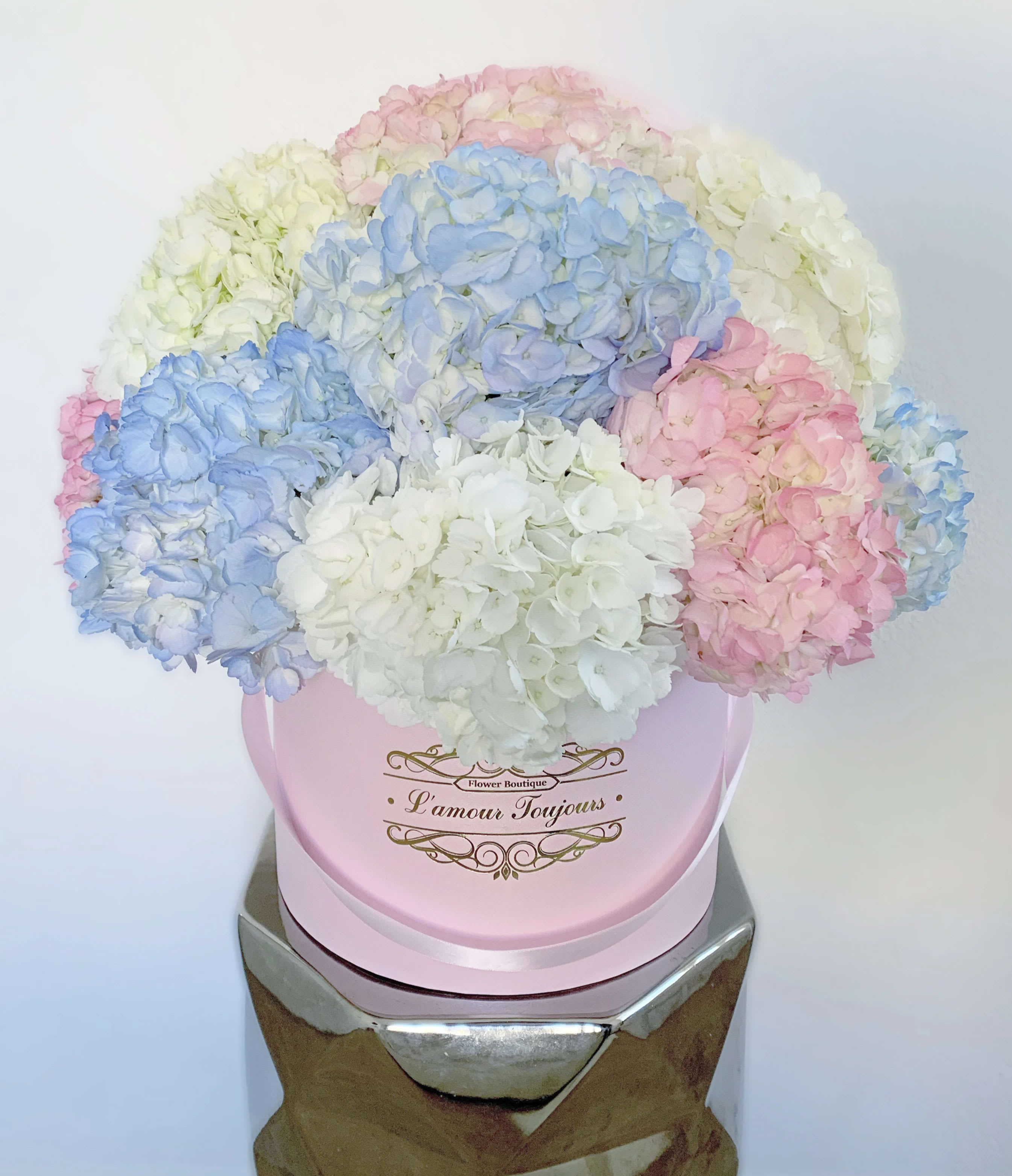 Pastel blue, pink, and white hydrangea arrangement in a pink hat box on a reflective pedestal