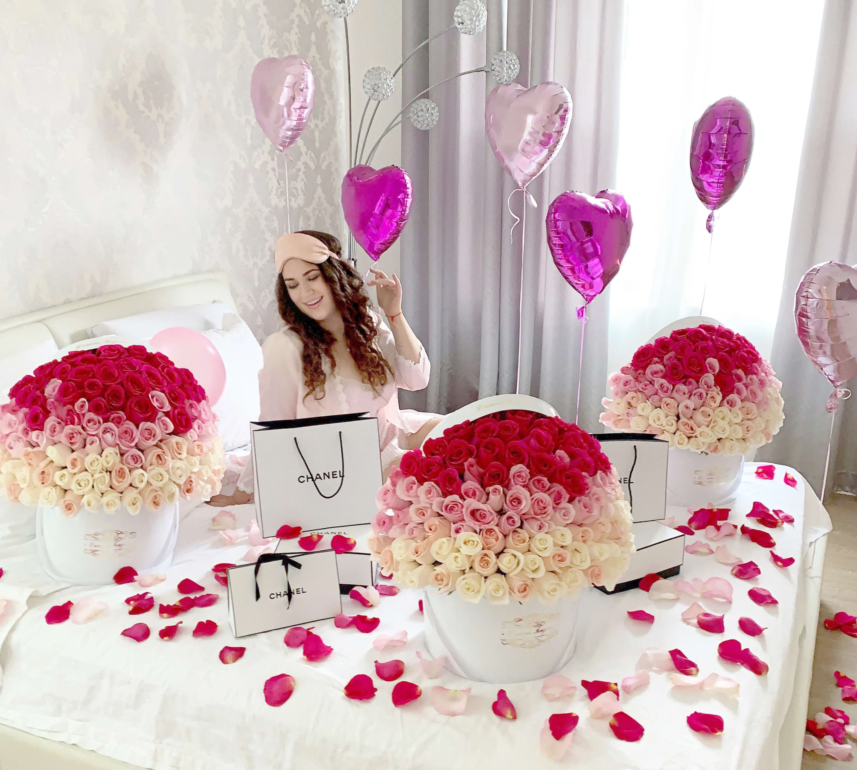 Large hatbox arrangements of pink and white roses on a bed with heart balloons and gift bags.