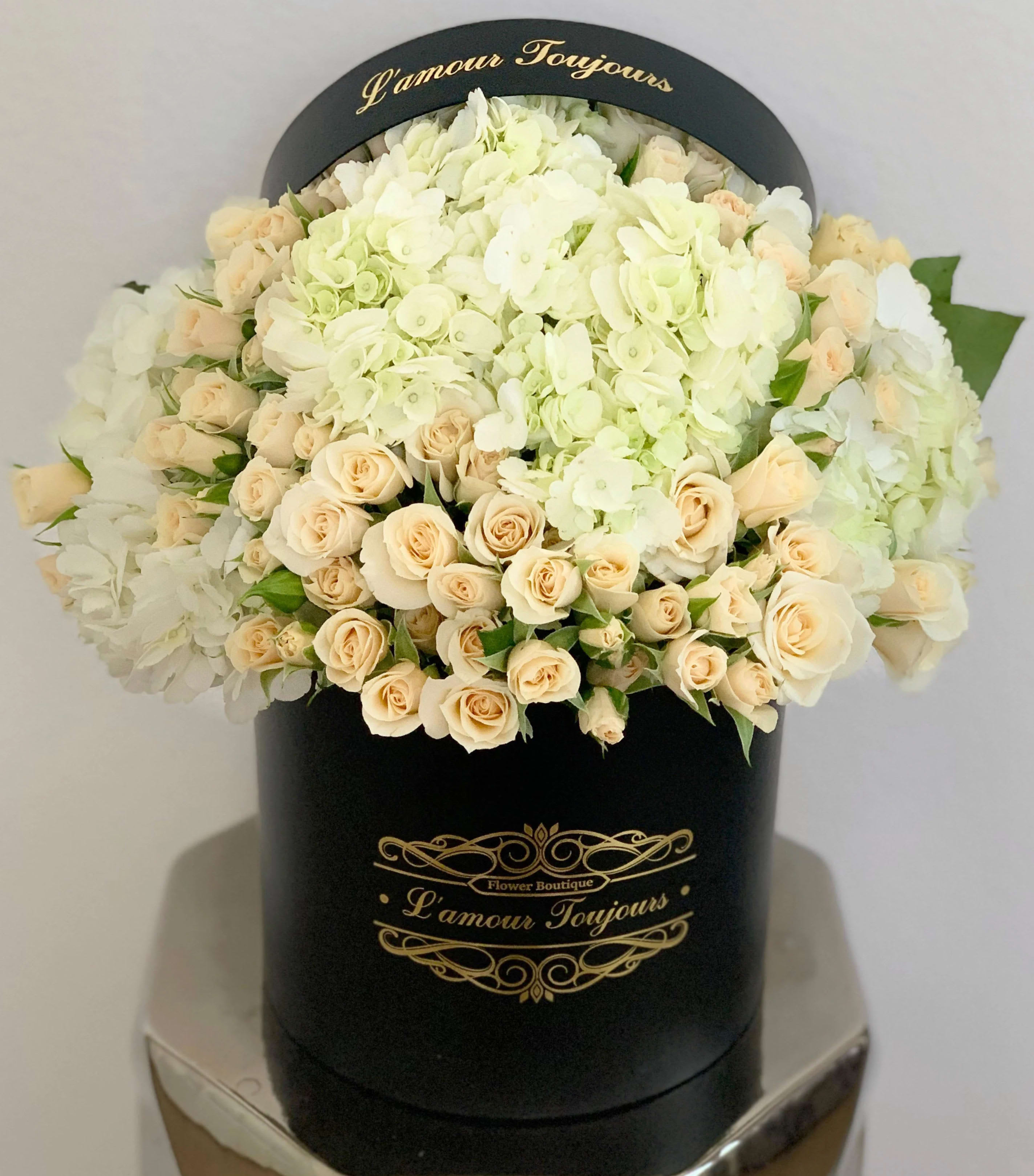 Round black hatbox filled with cream roses and white hydrangeas with gold L'amour Toujours lettering