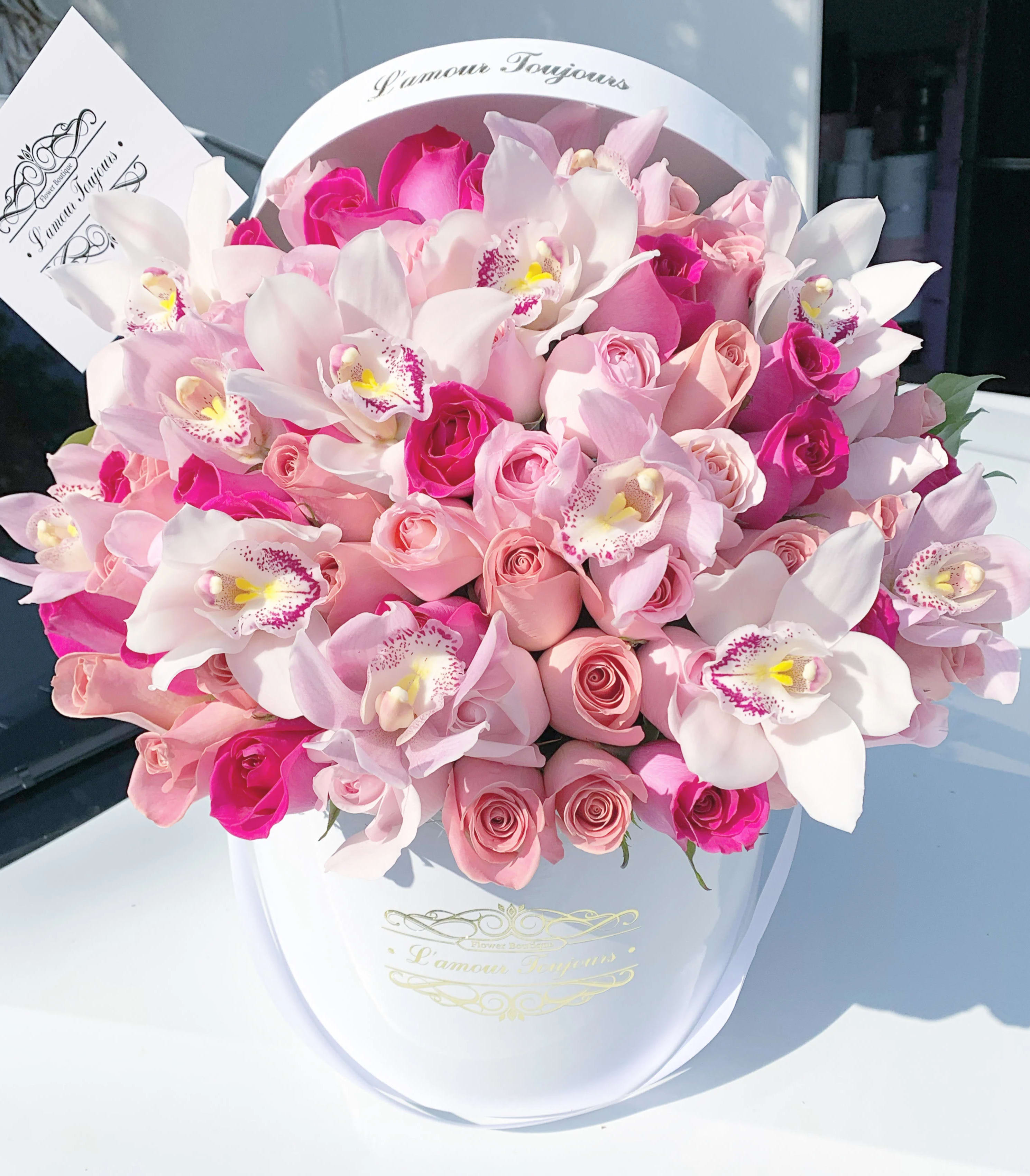 Hatbox arrangement of pink roses and white orchids in a white round box with a card.