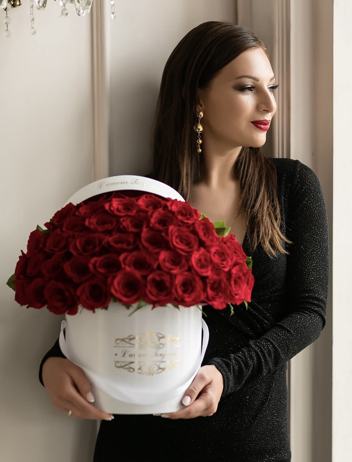Woman in a black dress holding a round white box filled with dozens of red roses.