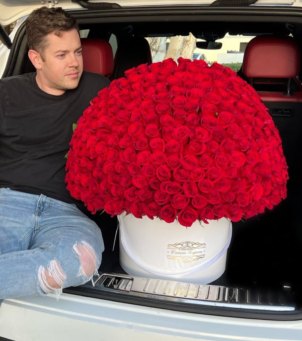 Large dome of red roses in a white hatbox in a car trunk beside a seated man