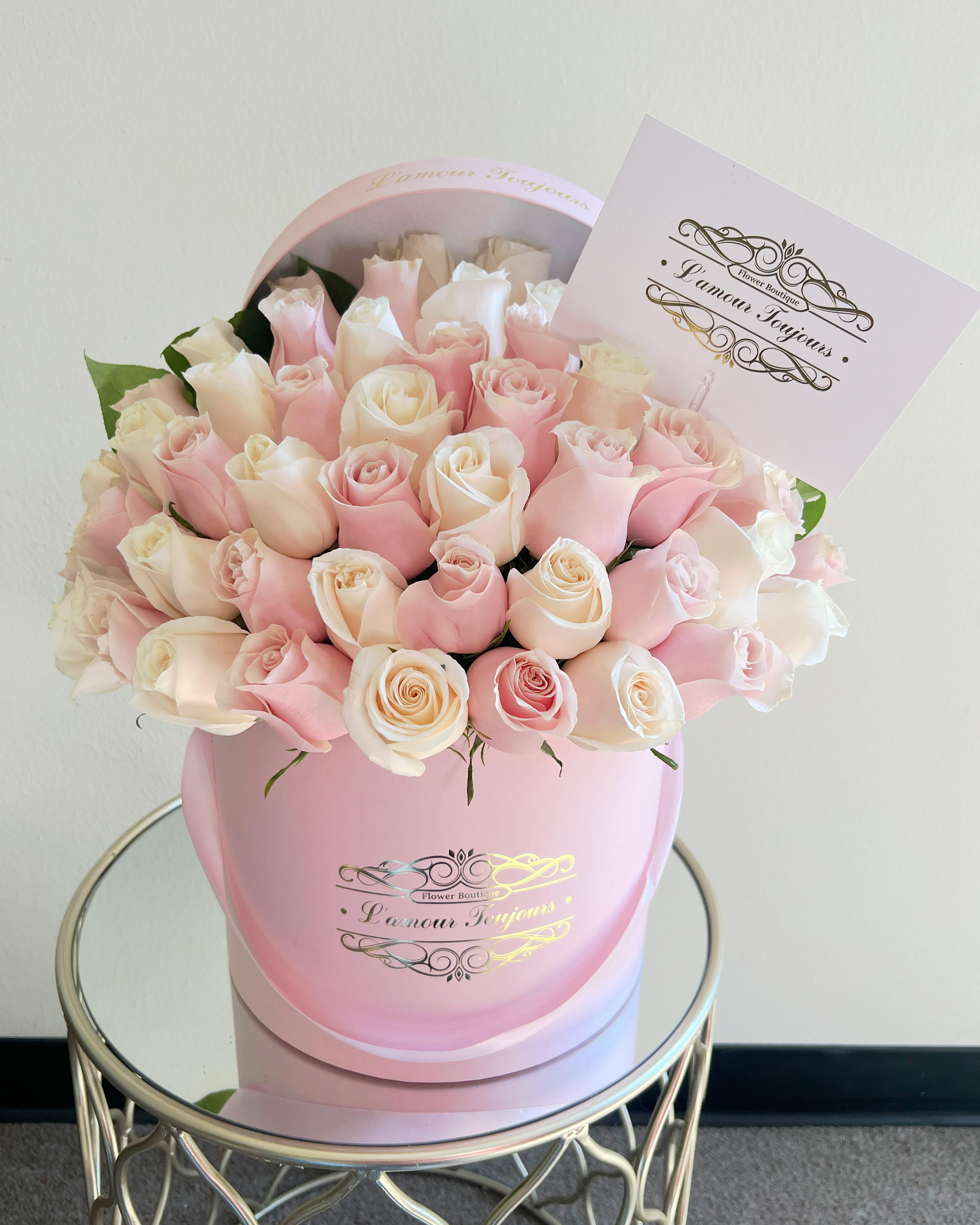 Round box of pale pink and cream roses with a card on a mirrored side table