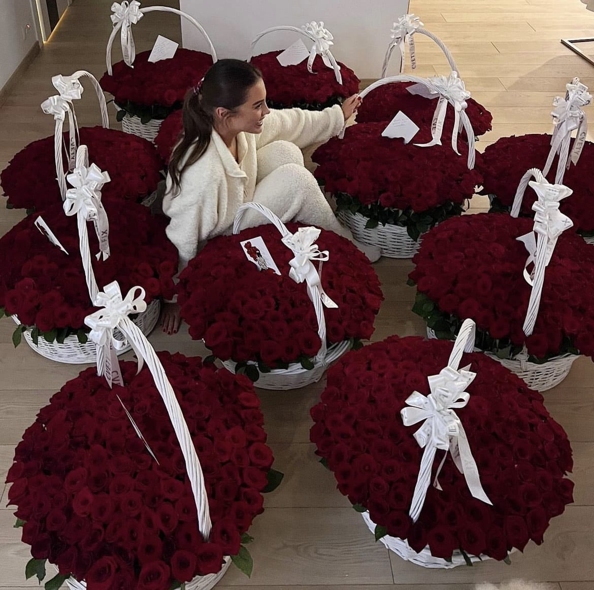 A woman sits among large white baskets overflowing with deep red roses and white bows.