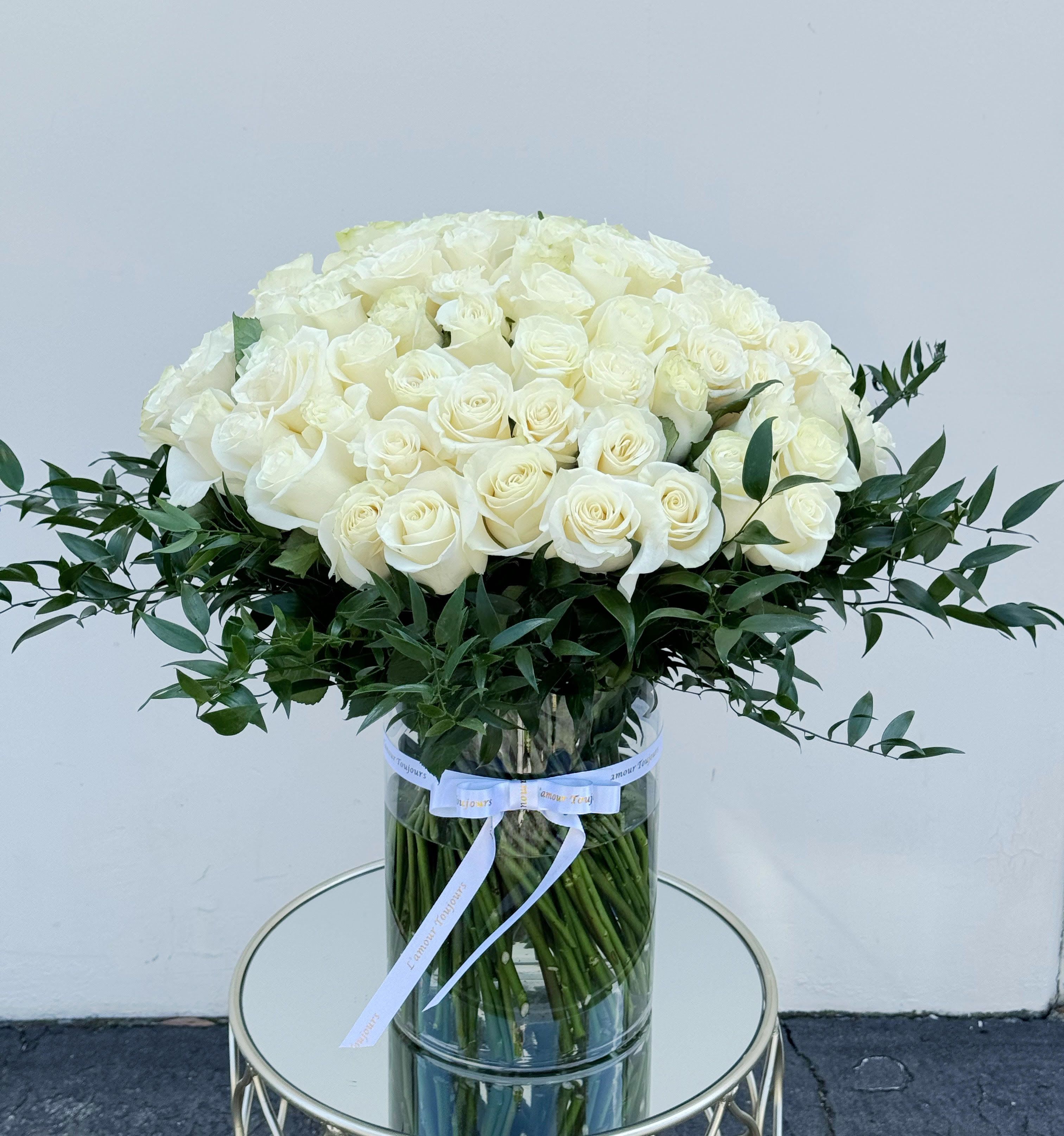 Large arrangement of cream roses with a collar of dark foliage in a tall clear cylinder vase.