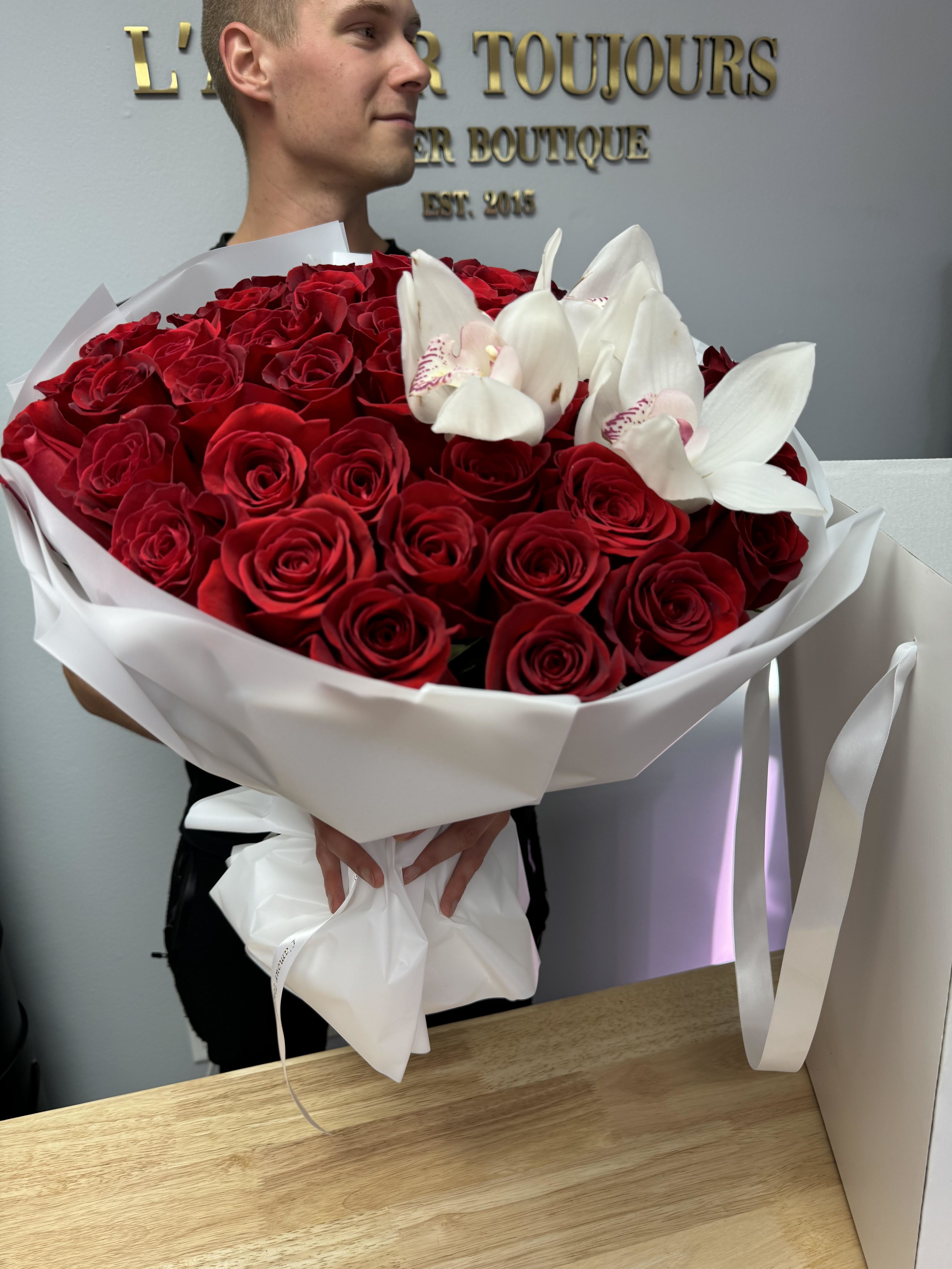 Large bouquet of red roses with white orchids wrapped in white paper, held by a person indoors