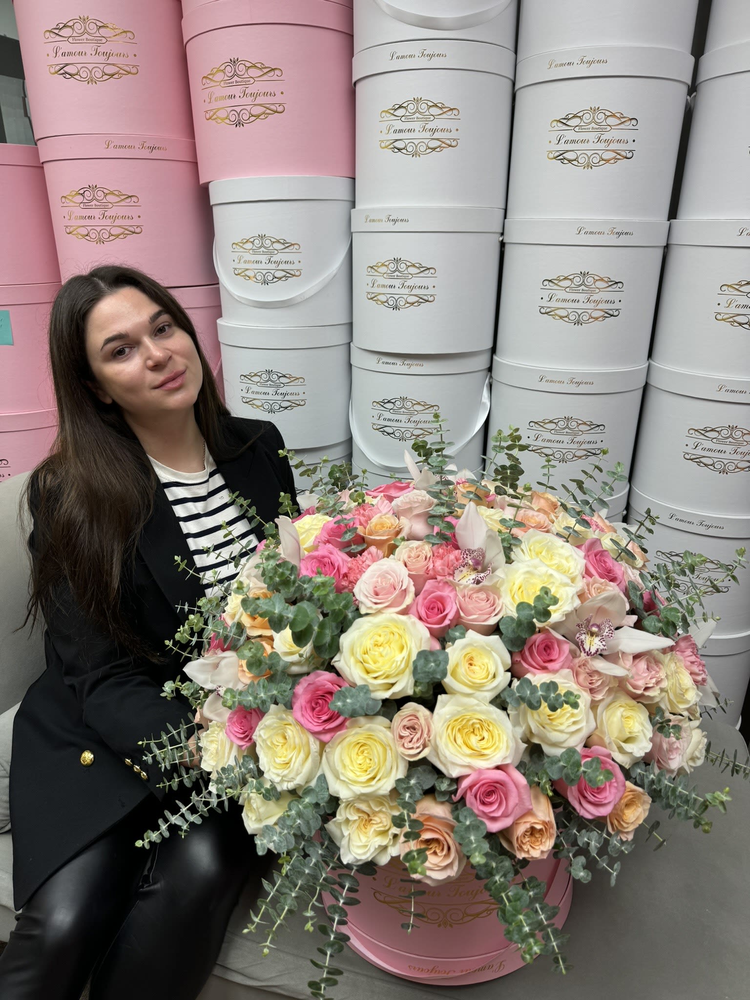 Large pink hatbox arrangement of pastel roses with orchids, next to a seated woman.