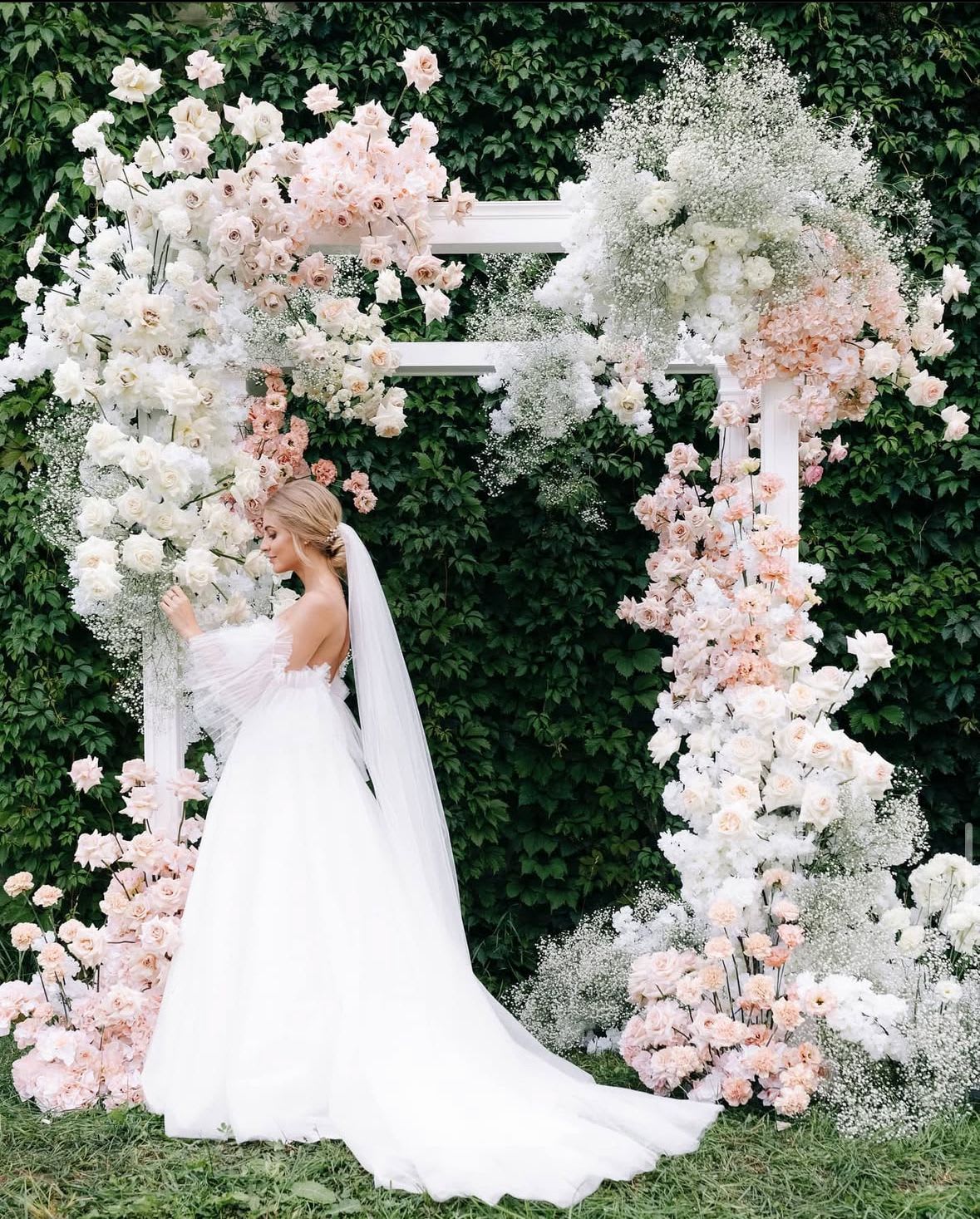 Bride in a white gown beside a floral wedding arch with white and blush flowers