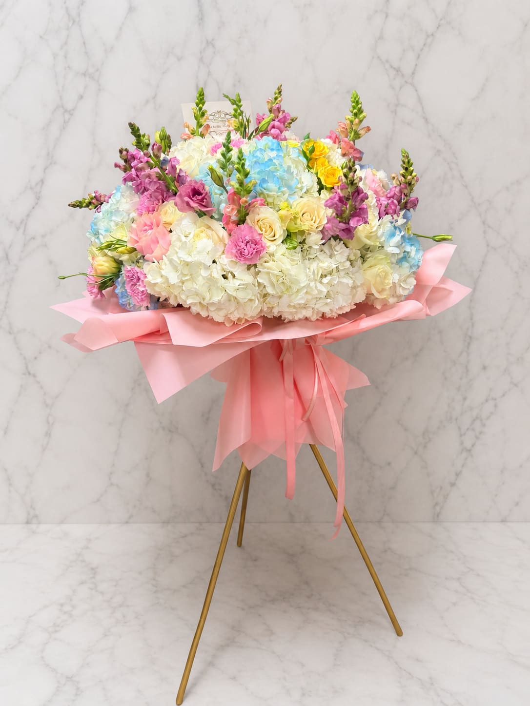 Standing Signature Bouquet  - A lush display of pastel blooms makes this standing bouquet an unforgettable way to celebrate-perfect as a statement piece, a birthday surprise, or a grand congratulatory arrangement. The arrangement type and occasion are clear from the first glance: a standing flower arrangement designed to mark special moments and let the recipient feel truly honored and celebrated.  Dense, soft white and pastel blue hydrangeas form the cloud-like base, while pink and cream roses add warm, romantic highlights. Upright purple and pink snapdragons give height and a graceful vertical rhythm, creating a layered composition that reads elegant and joyful. The bouquet arrives wrapped in pale pink paper with a flowing pink ribbon and is presented on a gleaming gold tripod stand for immediate impact in any entryway or event space.  This is the kind of floral gift that makes someone feel seen on a milestone day-whether for a wedding, anniversary, or big birthday. Sized to command attention, the arrangement is ready to display on the included gold stand; light misting of the blooms and keeping them away from direct heat will help maintain freshness for as long as possible.