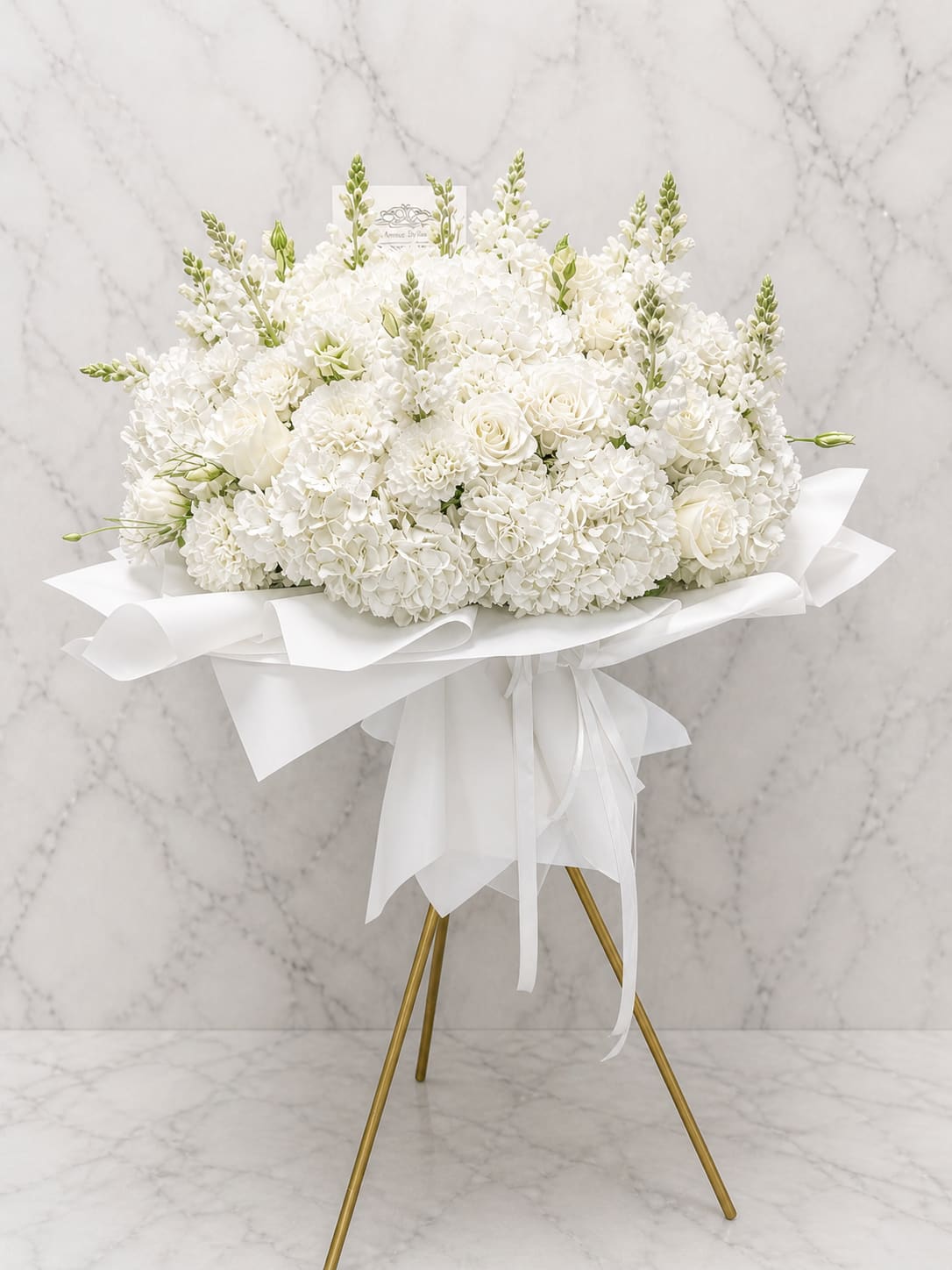 All-white standing signature bouquet - A serene all-white standing bouquet that makes a quiet, elegant statement for sympathy, memorial services, or a peaceful wedding display. The arrangement's dense, rounded mass of blooms is accented by taller upright spikes, creating a layered silhouette that reads graceful from every angle-perfect as a sympathy standing spray or a solemn tribute to mark a life and offer comfort.  Crafted from ivory roses, cloud-like hydrangeas, and fragrant stock, with small pale-green buds for a hint of fresh contrast, the textures move from soft clusters to tighter rose centers for visual depth. Wrapped in crisp white paper and tied with a flowing white ribbon, the bouquet is displayed on a gold tripod stand for a formal, refined presentation that suits chapels, funeral homes, or elegant ceremonies.  Designed to help the person you're honoring feel truly remembered, this arrangement sends a message of calm respect and heartfelt sympathy. Sizing and delivery options can be selected at checkout to ensure the standing spray fits the service space; minimal care is needed once placed on its stand-keep cool and out of direct sunlight for best longevity.