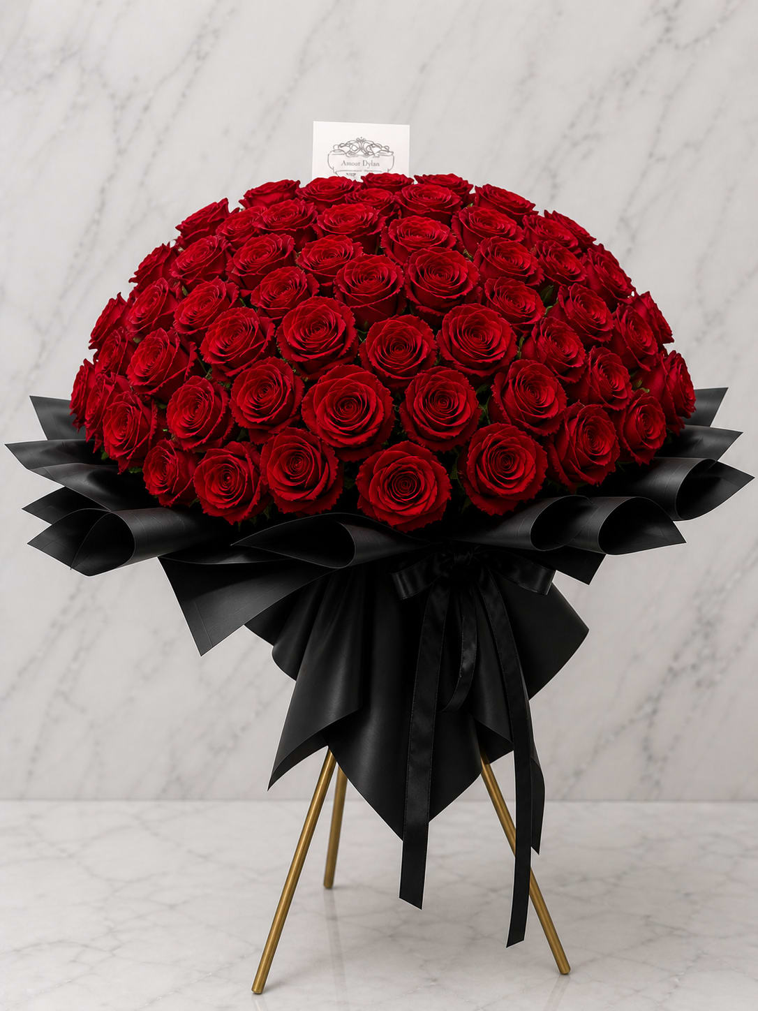 150 Red Roses Standing Bouquet - A dramatic cloud of 150 deep red roses creates an unmistakable statement-this red roses standing bouquet is designed to make someone feel profoundly adored. Perfect for an anniversary or a grand romantic gesture, the tightly packed, dome-shaped blooms give a lush, velvety visual that reads as pure romance the moment they enter the room.  The arrangement features a dense mass of vivid red roses wrapped in glossy black paper for a striking contrast, finished with an oversized bow and a small floral card tucked at the top. Mounted on a gold tripod stand, the bouquet's formal silhouette and luxe materials (black wrap, ribbon, and gold stand) elevate the presentation so it reads like a keepsake moment rather than a casual bouquet.  Made to mark a milestone or sweep someone off their feet, this standing bouquet is ideal for anniversaries, proposals, or any occasion where you want the recipient to feel deeply celebrated. Place away from direct heat and change the water if stems are kept in a vase; the floral card and ribbon complete the gift-ready display for immediate delivery and display.