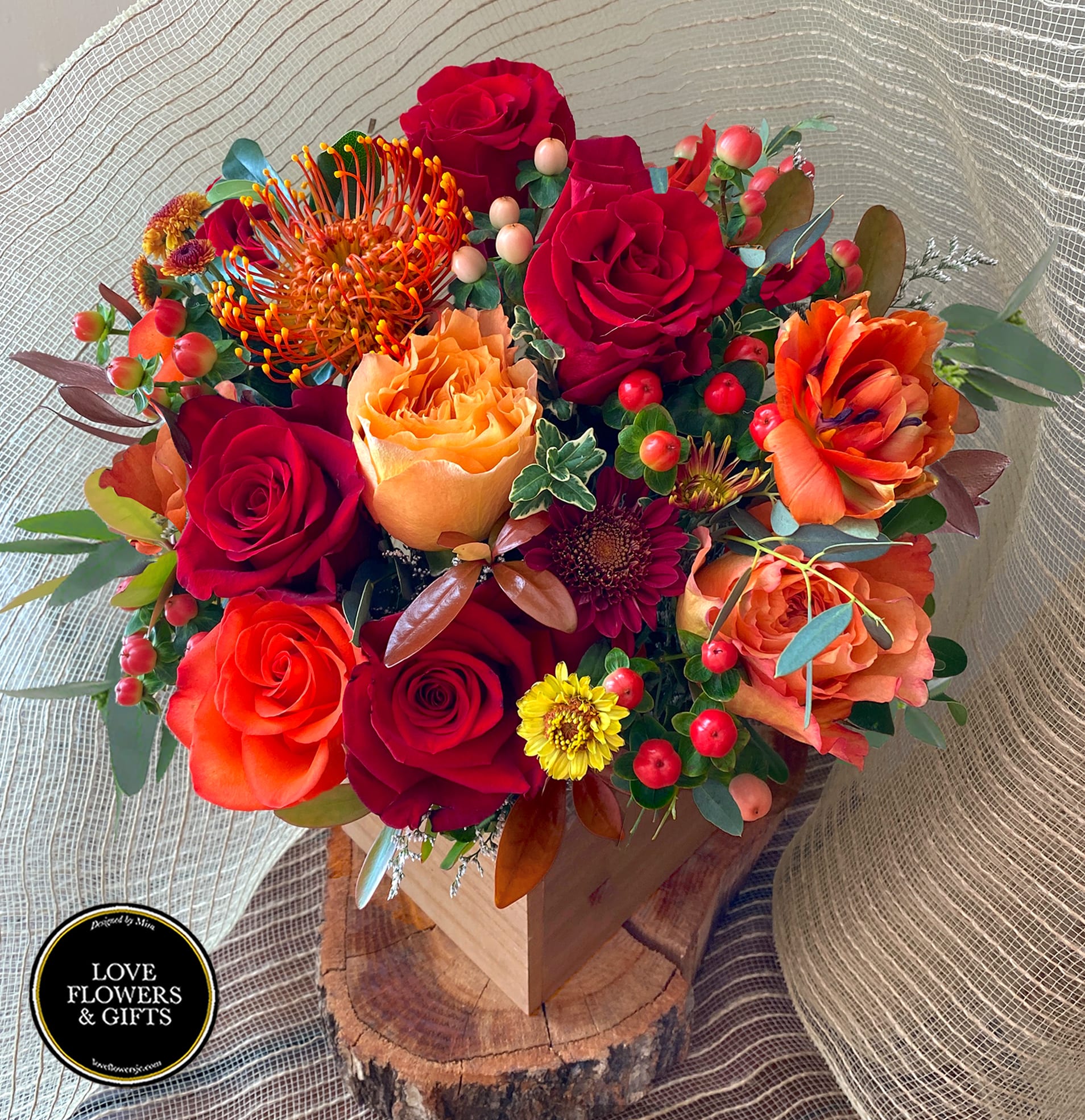 Red and orange rose arrangement with protea and yellow mums in a wooden box on a wood slice base