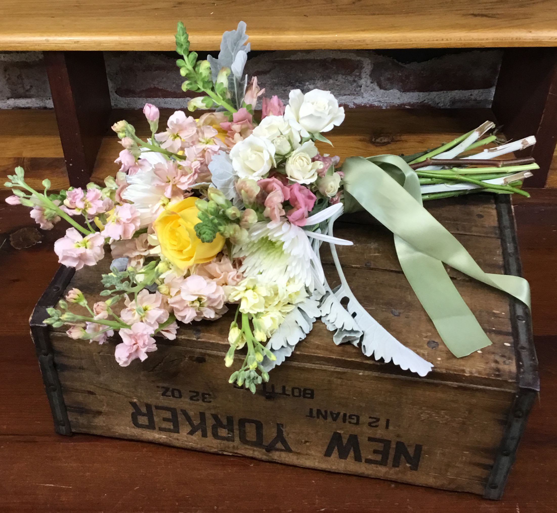 Loose hand-tied wedding bouquet of pastel blooms with a yellow rose and sage ribbon on a wooden crate.