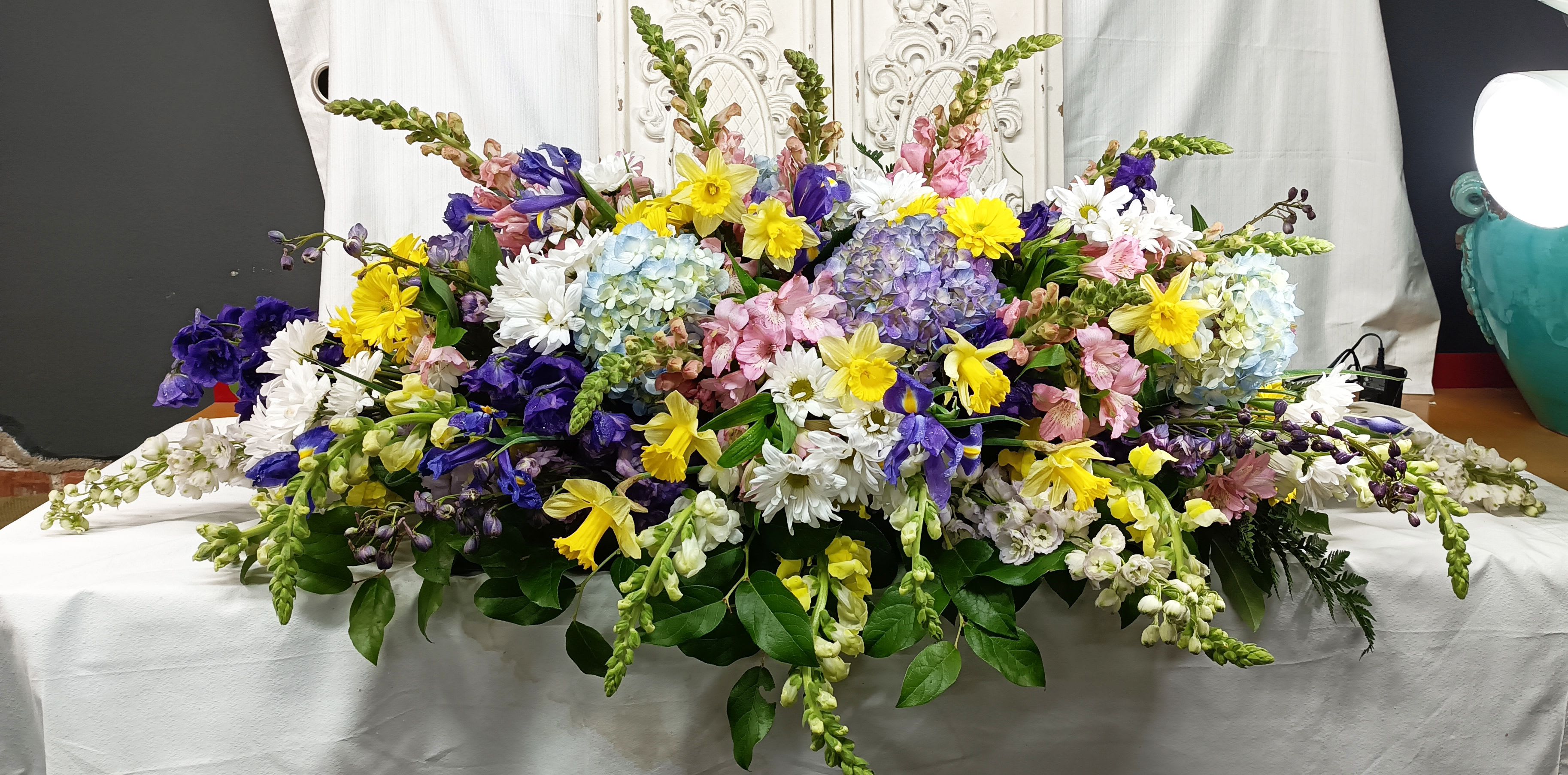 Spring Floral Casket Spray - A generous, low casket spray that balances dramatic red roses and stargazer-style lilies with softer pinks, whites and fresh spring hues-designed for sympathy and funeral settings where a meaningful, elegant tribute is needed. The arrangement reads formal and expressive at the front of a service, yet its garden-like mix of colors also conveys warmth and comfort to the person you're honoring.  This spray features a dense, layered composition of roses, lilies, carnations, snapdragon-like spikes, hydrangea clusters, daisies, daffodil forms and iris accents woven with verdant greenery. Strong red accents sit alongside pale pinks, crisp whites, lavender and yellow for a full, abundant texture that spreads broadly across the casket surface. The low, sprawling shape creates a polished, substantial presentation while airy filler keeps the design feeling tender rather than heavy.  Appropriate for memorial services or to express condolences, this piece makes the recipient feel deeply remembered and cared for in a solemn moment. Available in a sizeable format to cover the casket's front, care is simple-kept cool and out of direct sun at the service to preserve freshness-please contact us for sizing options or delivery arrangements to ensure timely placement.