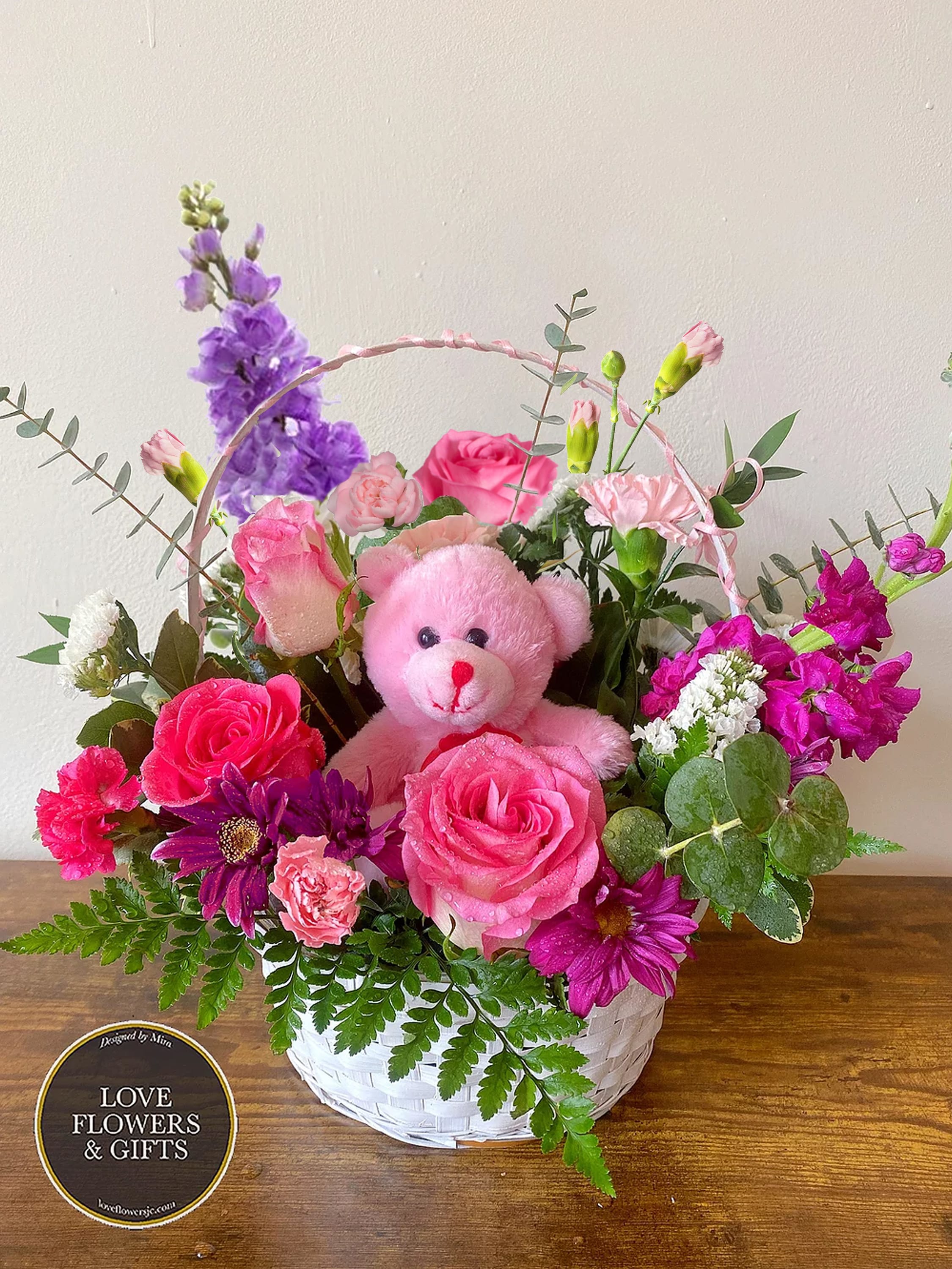 White basket of pink and purple flowers with a pink teddy bear in the center