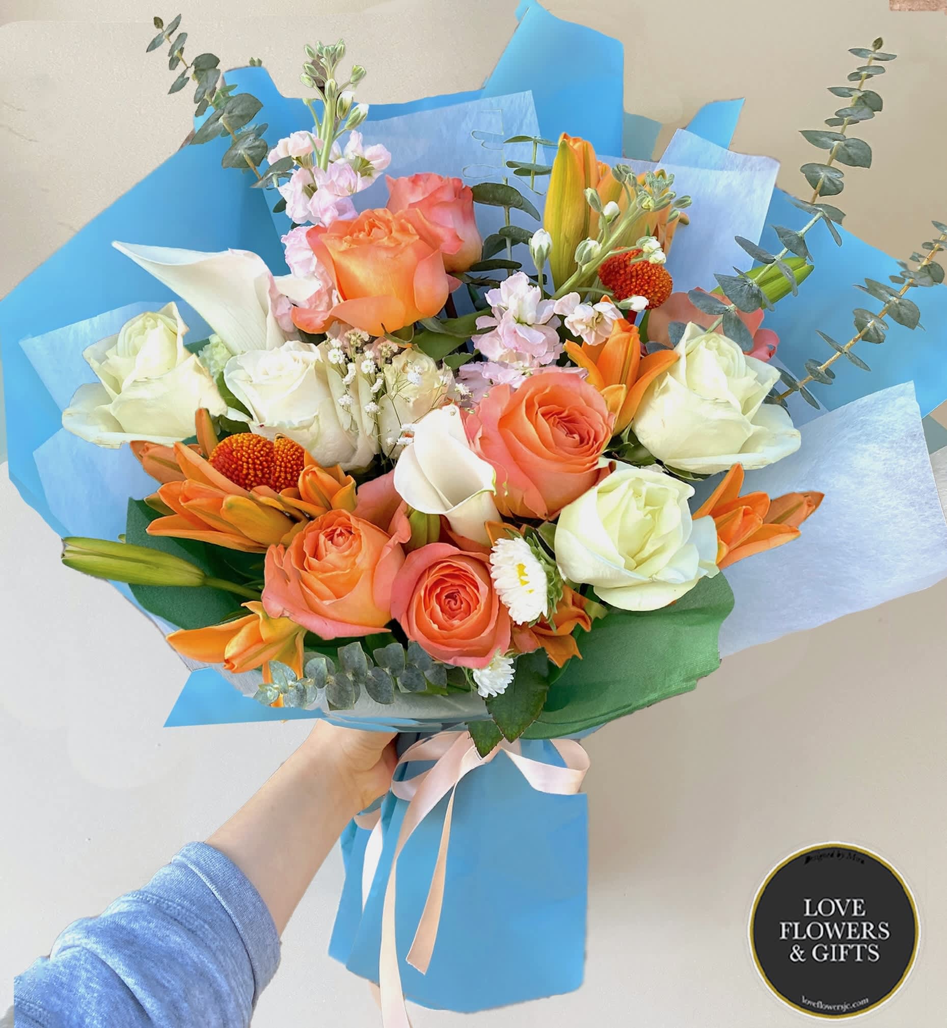 Hand-tied bouquet of peach and white roses with orange lilies and white blooms in blue wrapping.
