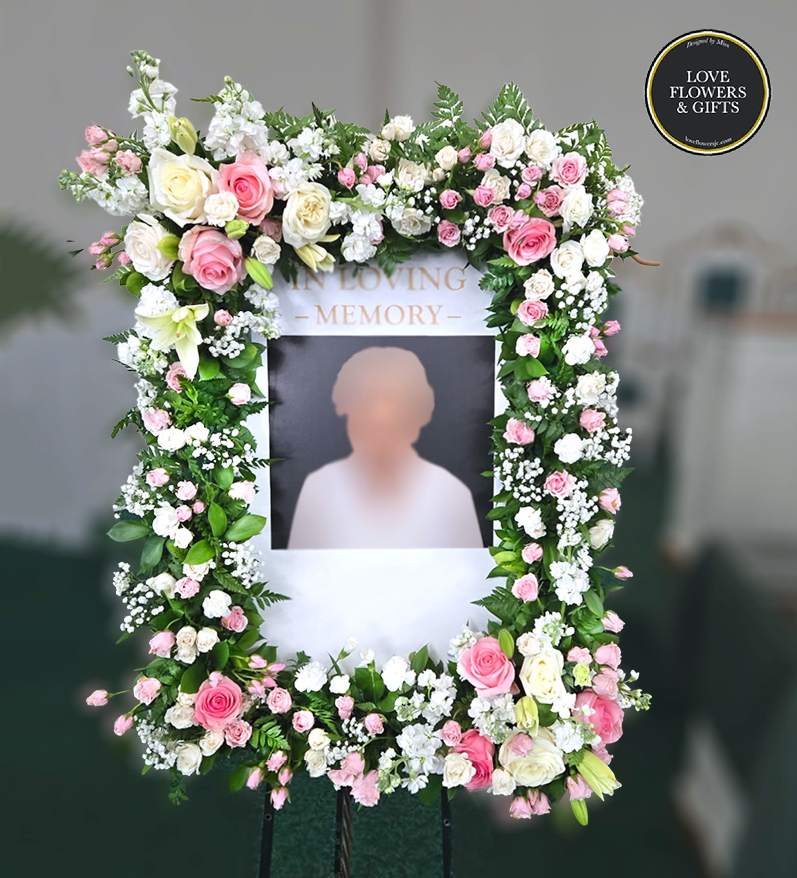 Tall rectangular memorial frame on an easel, bordered with pink and white flowers around a photo area.